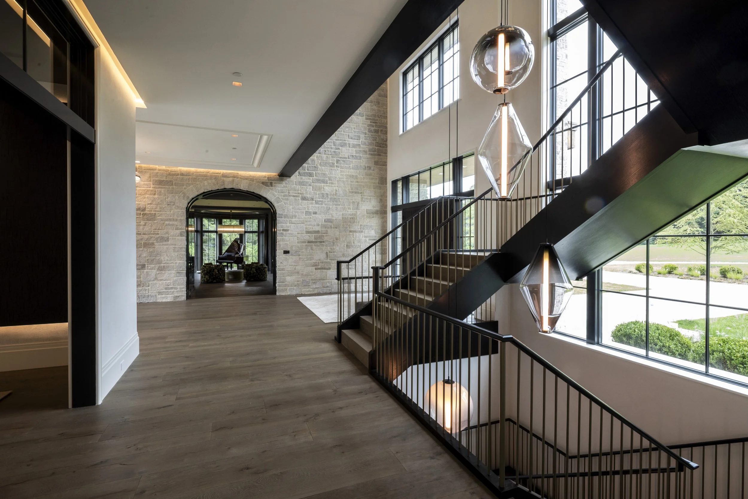 Modern interior of a spacious house with a staircase, large windows, pendant lights, and a living area with a grand piano in the background.