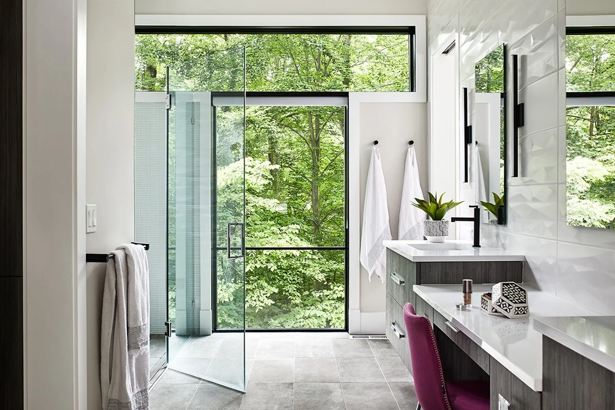 Modern bathroom with large glass shower door, white walls, gray floor tiles, and a vanity with a sink, mirror, and decorative items. Green trees are visible through the windows.