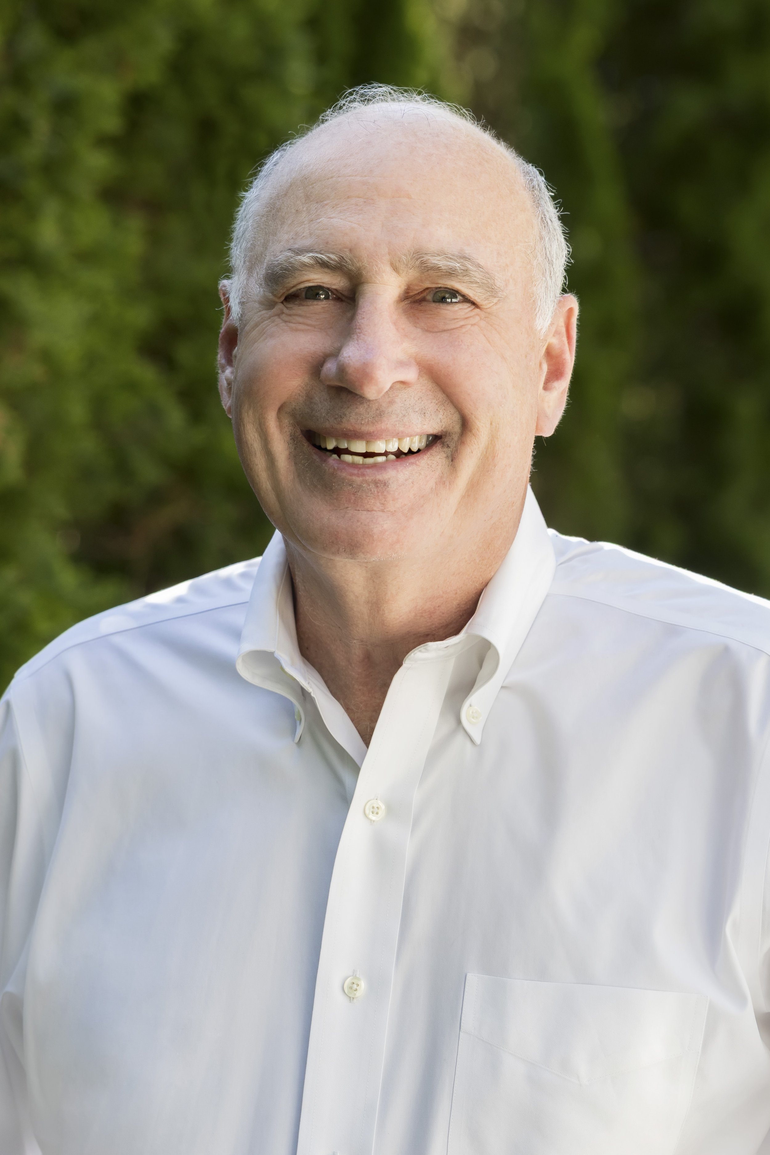 A smiling middle-aged man with short gray hair wearing a white dress shirt outdoors with greenery in the background.