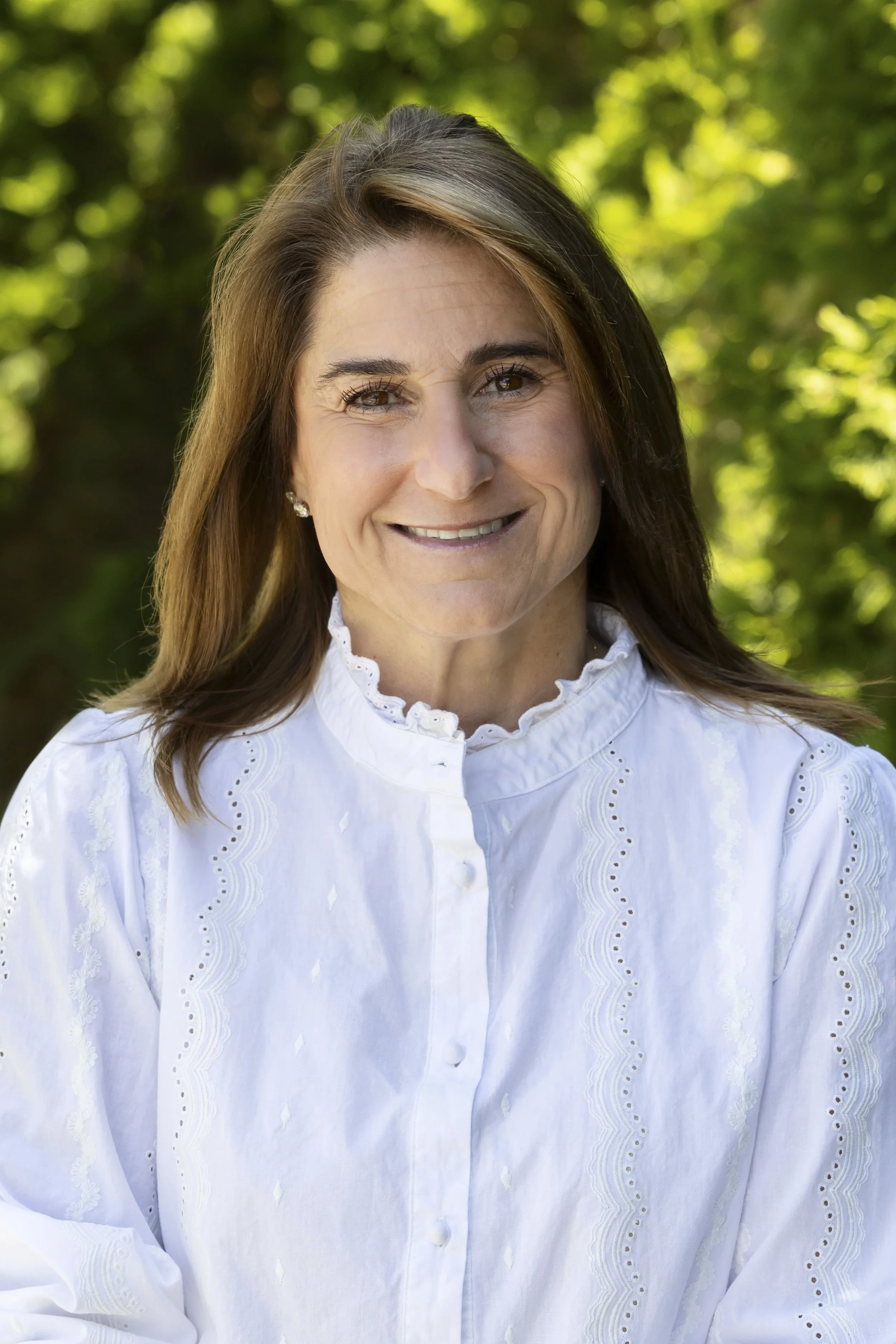 A woman with brown hair and light skin smiling outdoors, wearing a white embroidered blouse with a high ruffled collar, and earrings, with green blurred trees in the background.