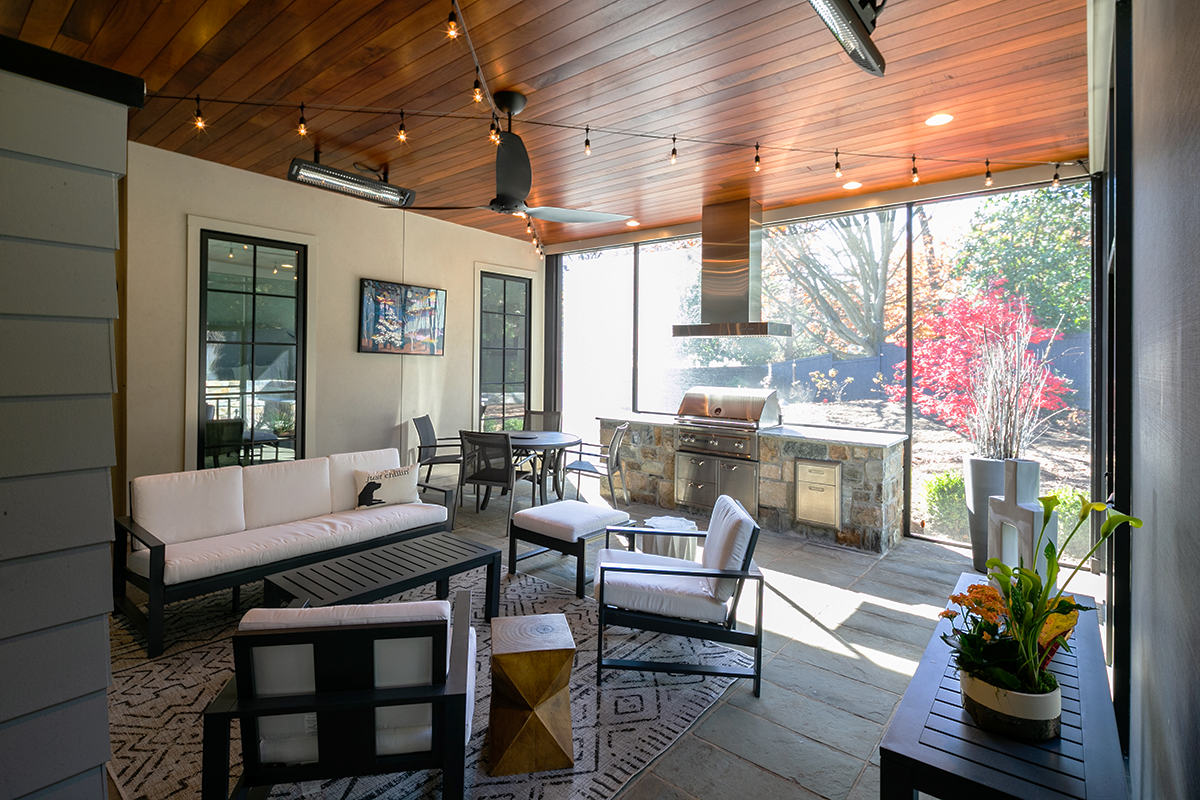 Indoor patio with lounge seating, a dining table, a kitchen grill, large windows, decorative plants, and string lights.