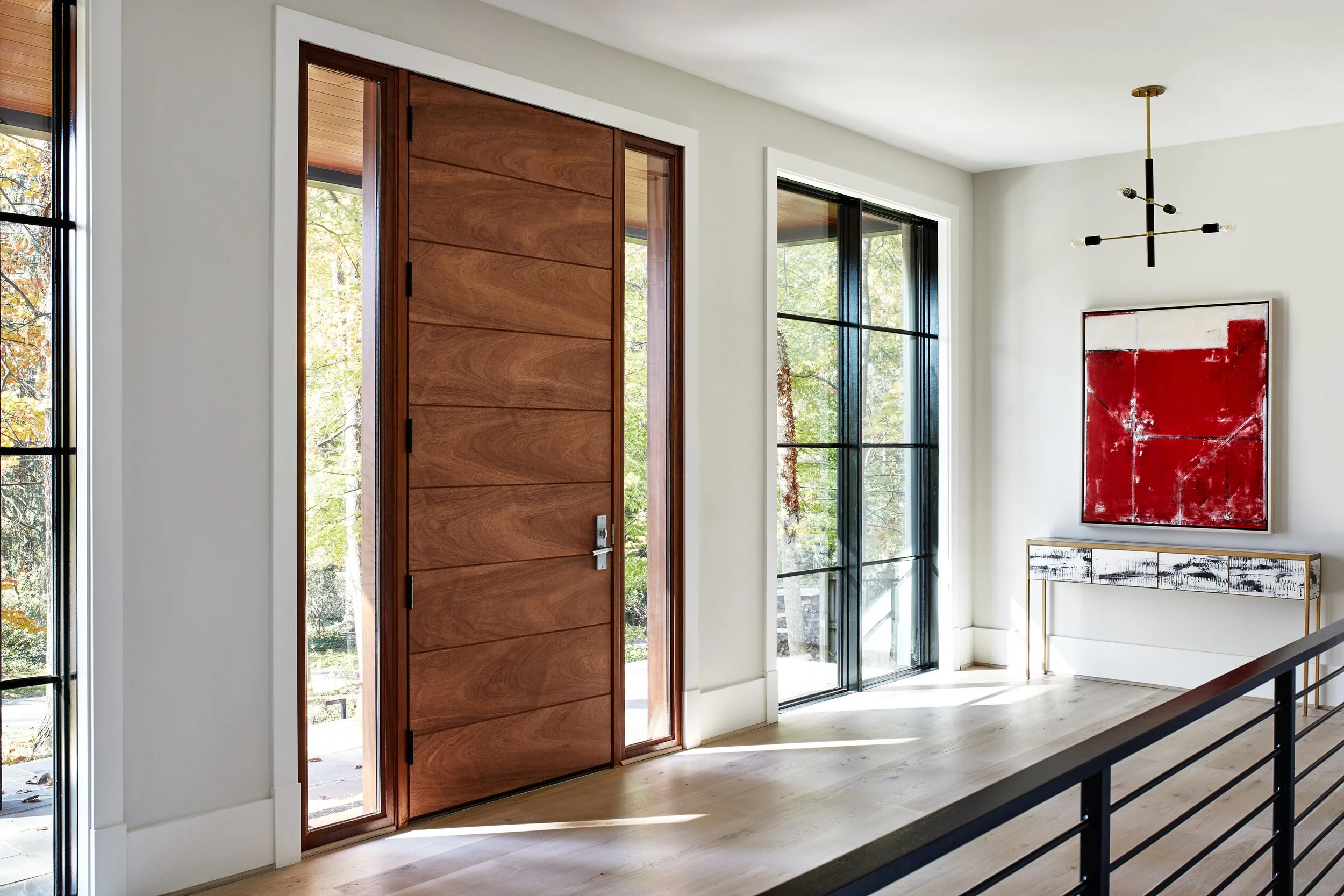 Modern interior with a wooden door with horizontal panels, large windows with black frames, and abstract artwork on the wall, featuring red and white colors. A minimalist chandelier hangs from the ceiling, and there is natural light coming through th
