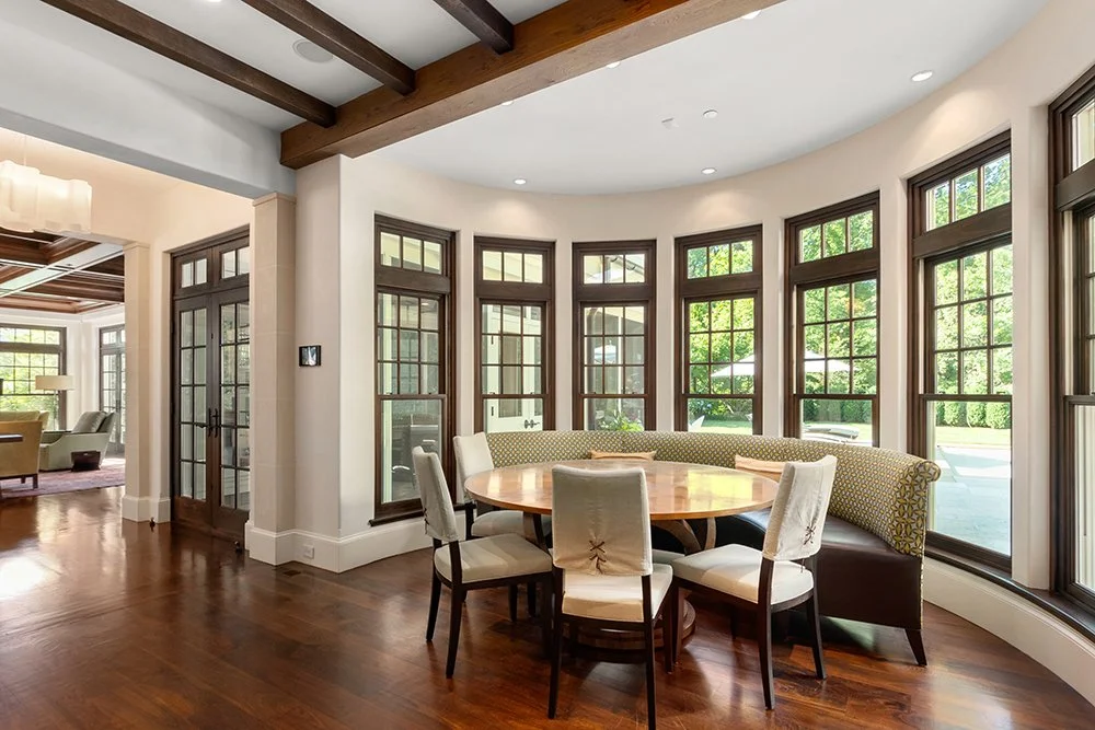 A dining area with a round wooden table, six chairs, and a curved window with multiple panes and brown trim, overlooking a green outdoor space.