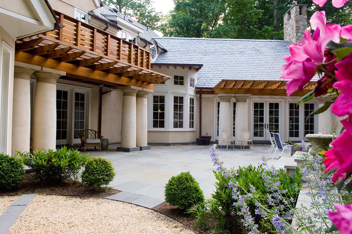 Spacious stone patio of a house with white French doors and large windows. The house has a wooden balcony and a shingled rooftop with trees and pink flowers.