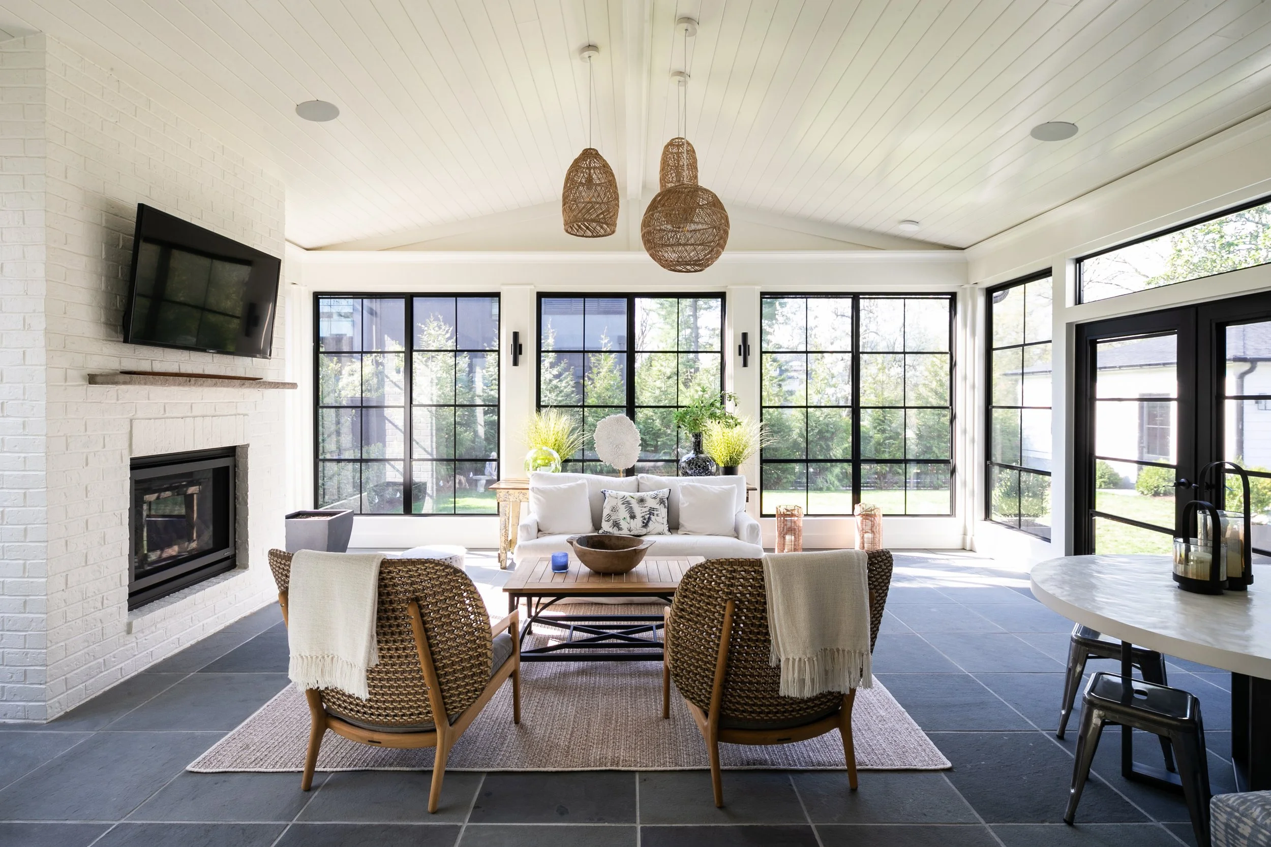 Bright sunroom with large windows, a white couch, wicker chairs around a wooden table, hanging woven pendant lights, a fireplace, and a mounted TV.
