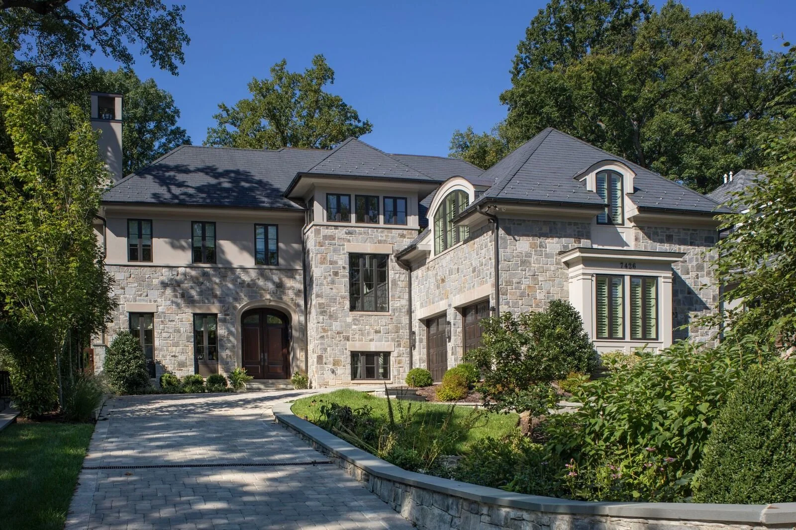 Large house with stone and stucco exterior, multiple windows including arched and rectangular designs, gabled roof, front driveway, manicured lawn, trees, and shrubs surrounding the property.