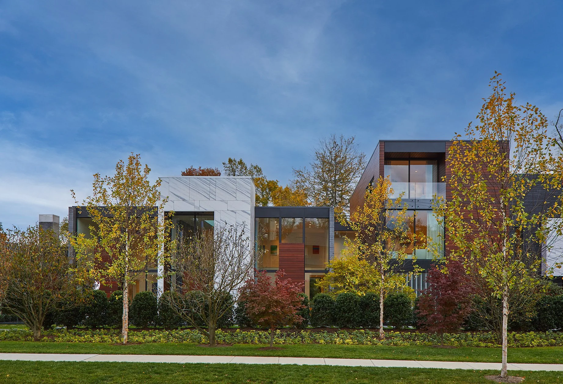 Modern multi-story residential building with large windows, surrounded by trees with autumn foliage and a well-maintained lawn under a partly cloudy sky.