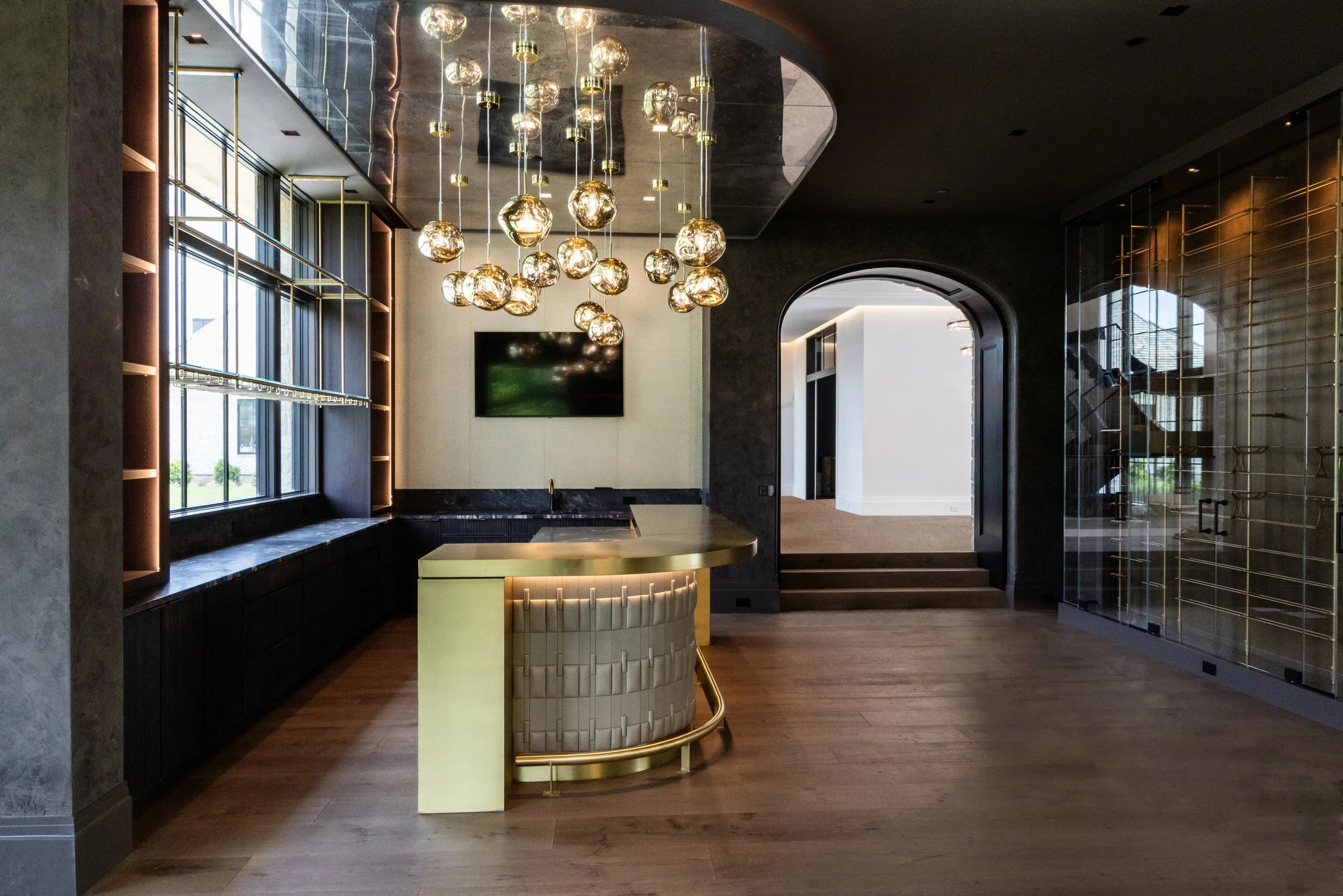 A luxurious interior with a modern chandelier composed of glass orbs hanging from the ceiling. The room has large windows on the left, framed with dark metal, and an arched doorway on the right. There is a green and gold curved counter in the center,