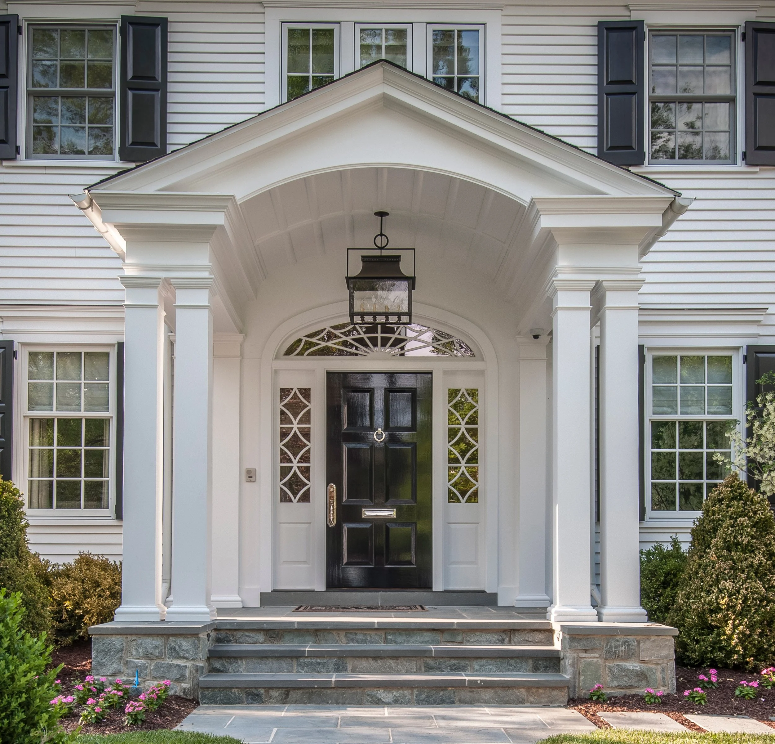 Front porch of a house with a black door, white columns, a gabled roof over the entrance, black window shutters, and a hanging lantern.