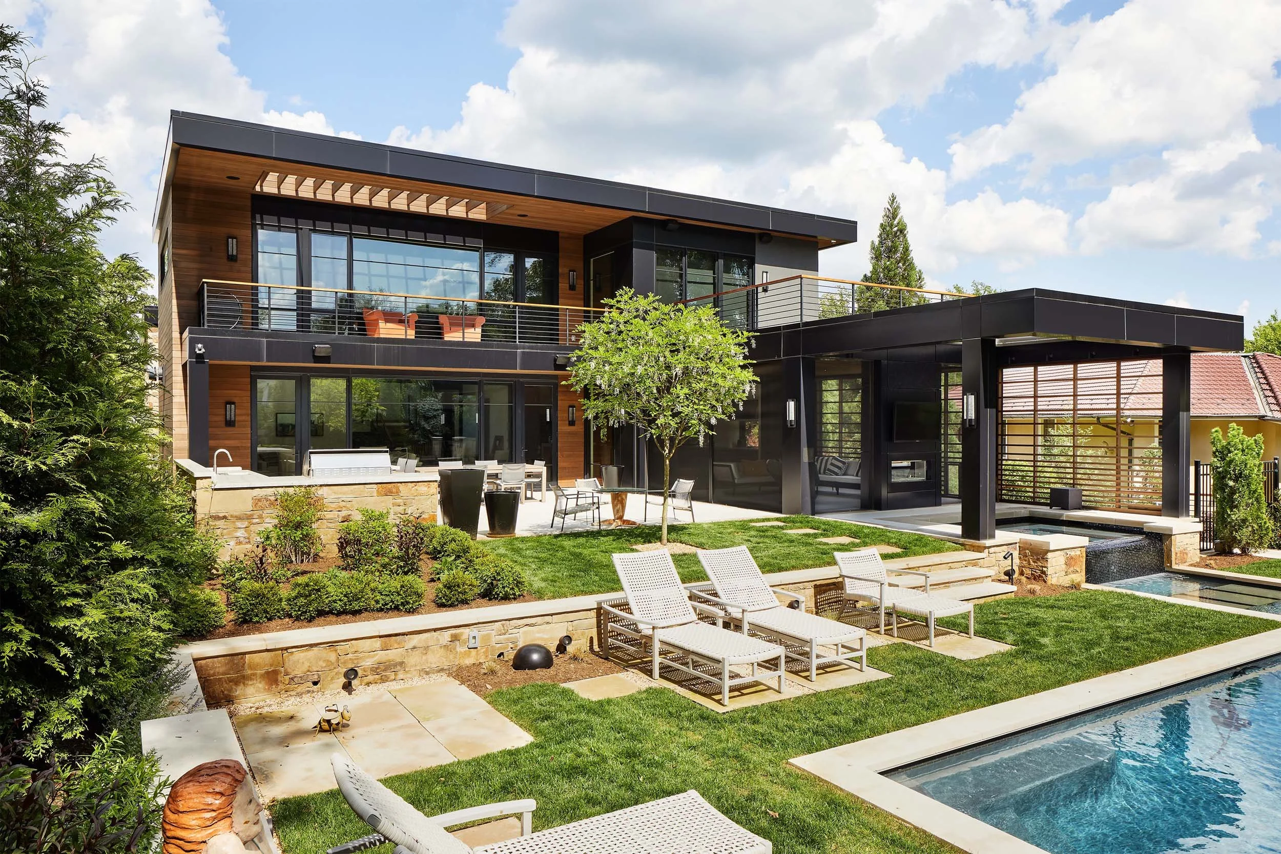 Modern two-story house with large glass windows, a balcony, and an outdoor pool area with lounge chairs surrounded by greenery and a bright, partly cloudy sky.