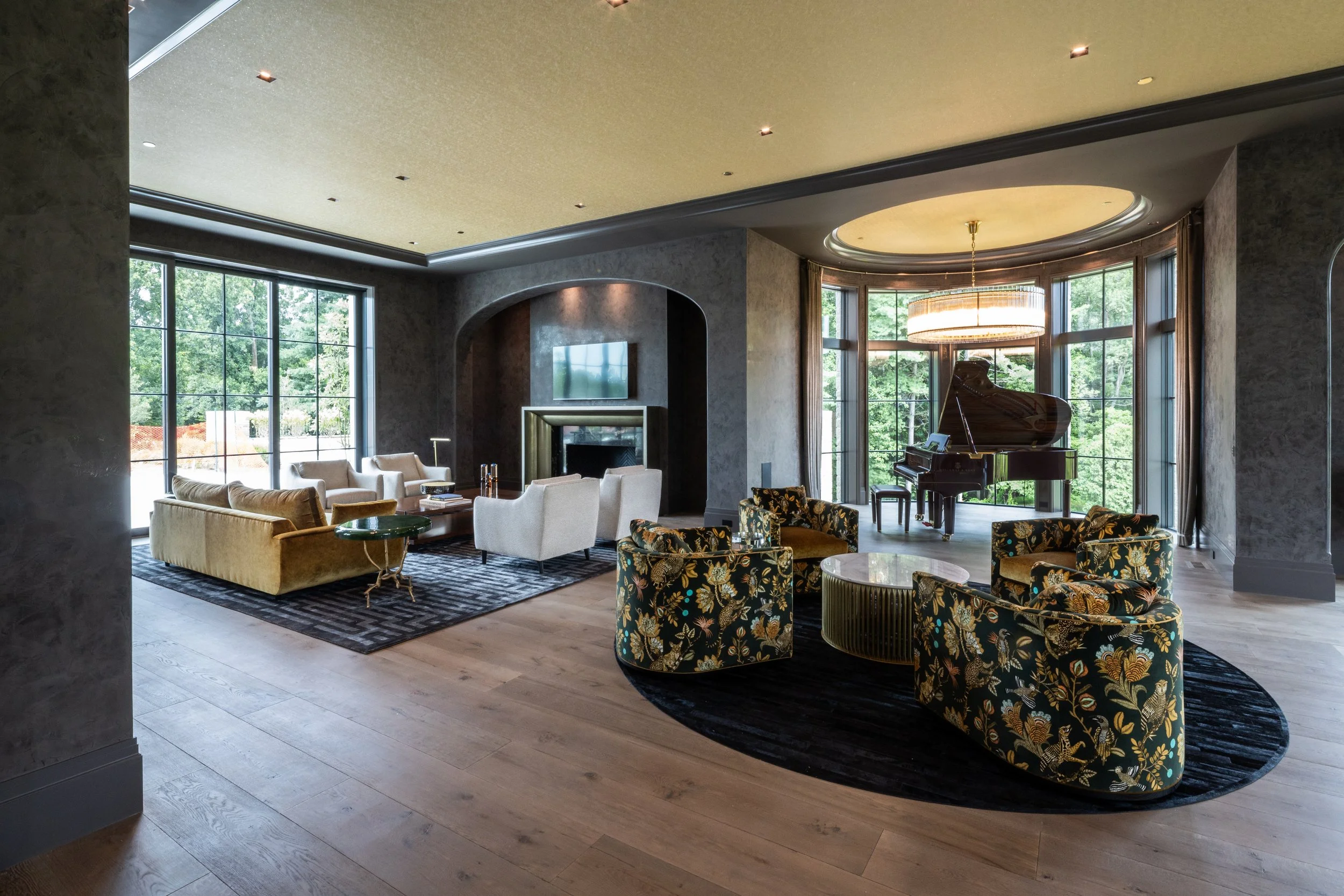 Luxurious, moody living room with large windows showing greenery outside, featuring beige sofas, floral armchairs, a grand piano, a fireplace, and a modern chandelier.