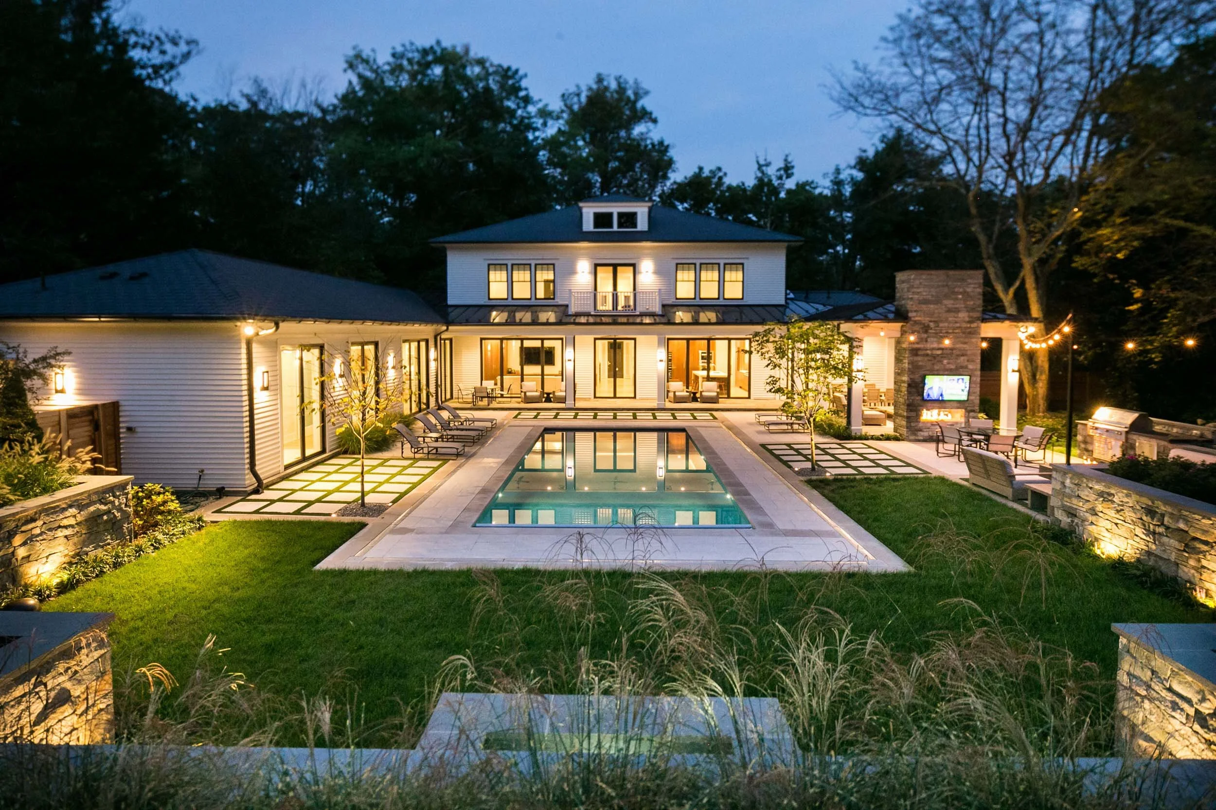 Nighttime view of a modern home with a lit outdoor pool, patio seating, and string lights, surrounded by trees.