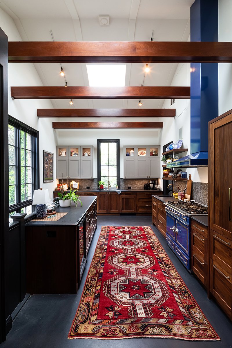 A spacious kitchen with wooden beams on the ceiling, gray and dark wood cabinetry, a red patterned rug, a black countertop, and large windows with black frames letting in natural light.