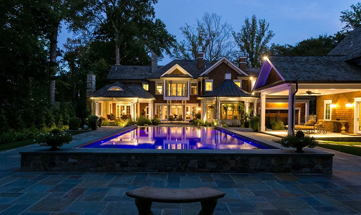 Large house with illuminated windows and a backyard swimming pool at dusk, surrounded by trees and outdoor seating areas.