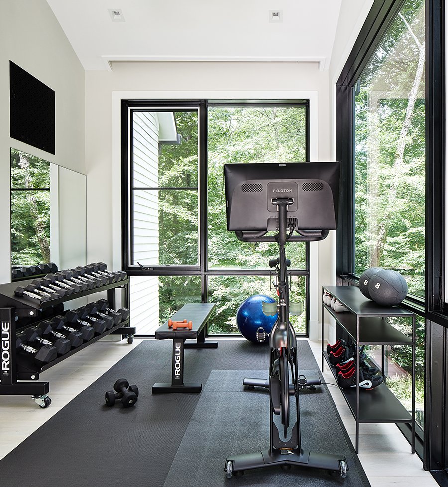A modern home gym with a stationary exercise bike facing large windows, a rack of dumbbells, a bench, dumbbells on the floor, a stability ball, medicine balls, and boxing shoes on a shelf.