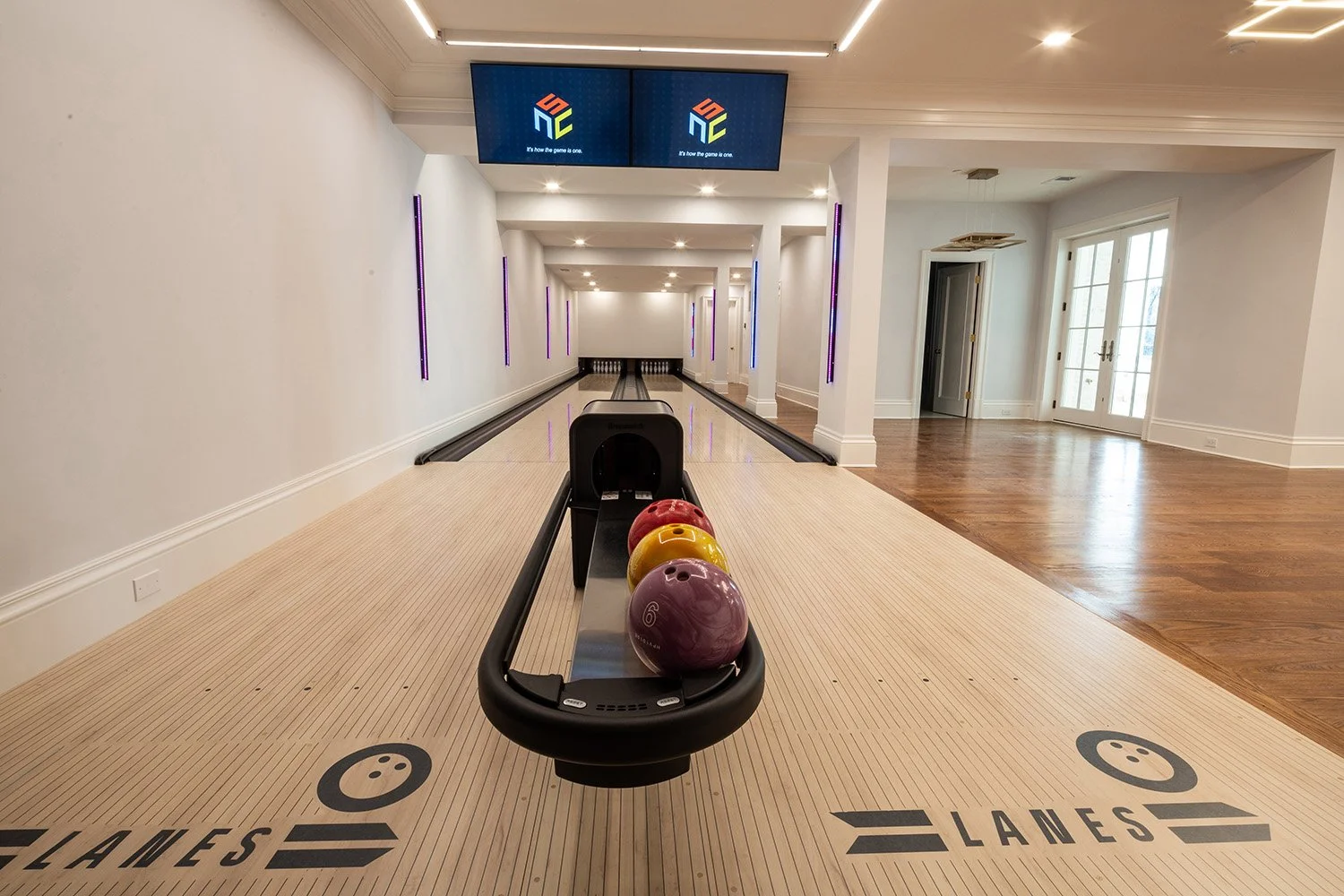 Indoor bowling alley with four colorful bowling balls on the ball return, lanes marked with 'LANES' and smiling face icons, and digital screens displaying a colorful logo and text in the background.