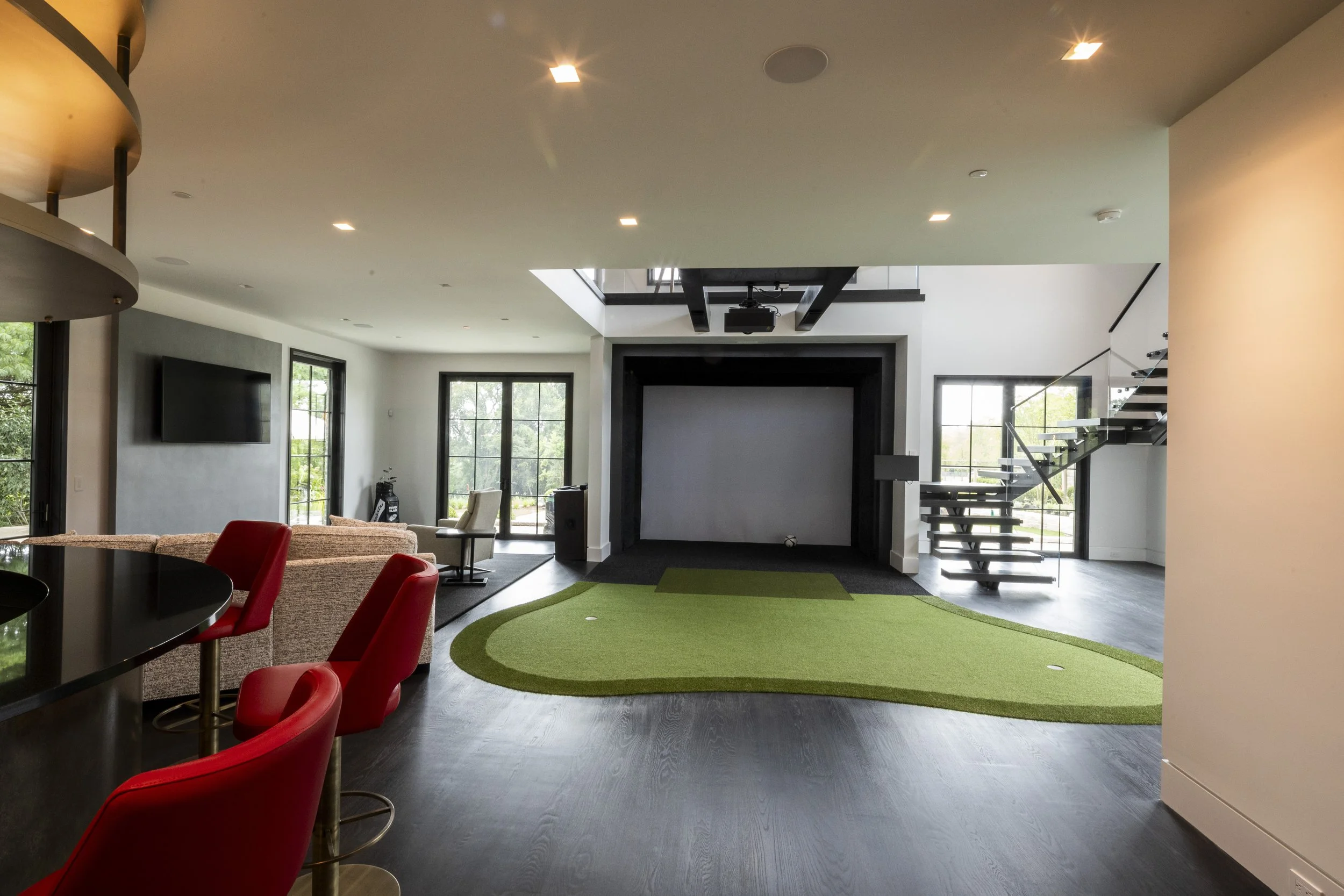 Modern living room with a large projector screen, green carpet, staircase with glass and black metal railing, windows with greenery outside, and contemporary furniture.