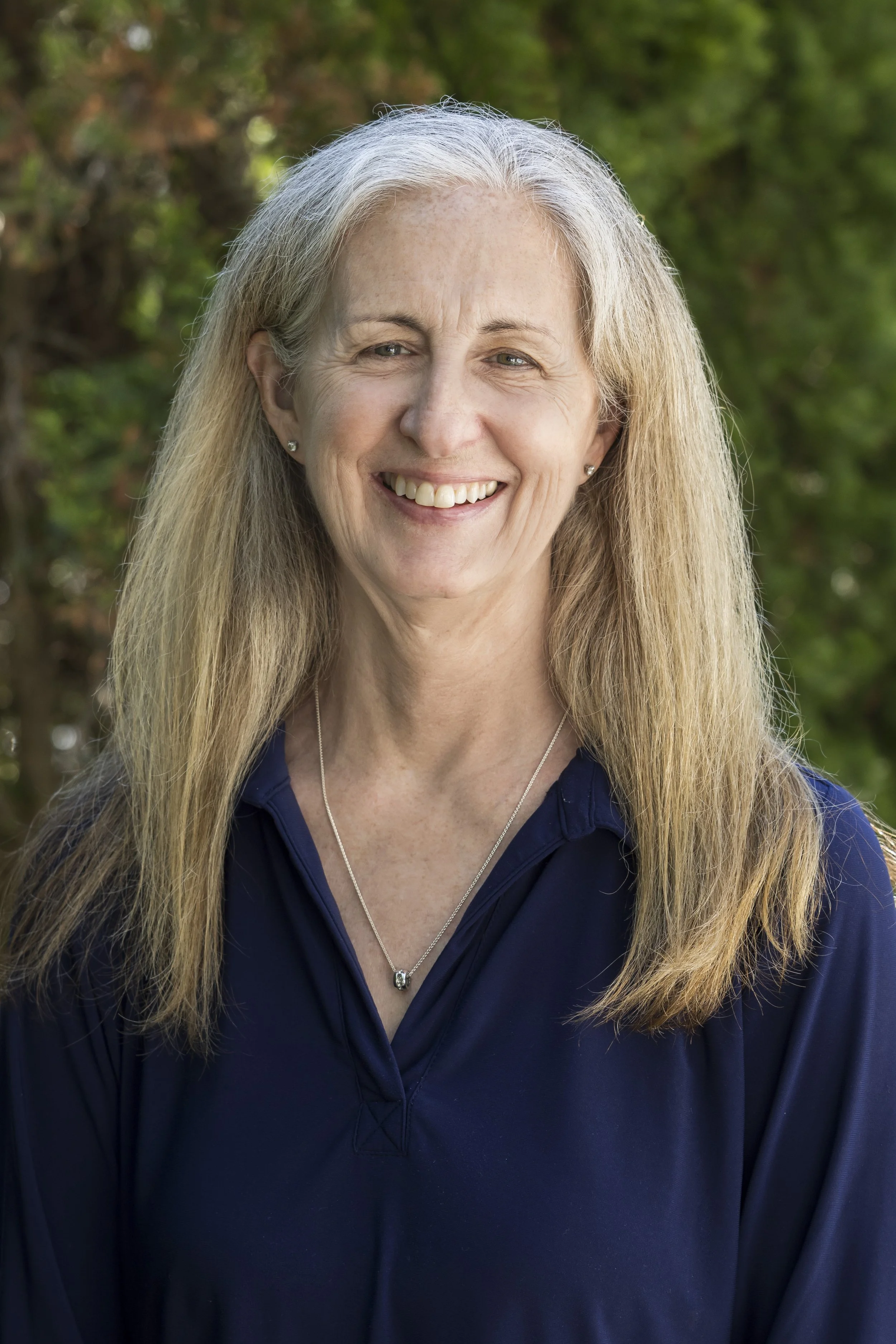 Close-up of a smiling middle-aged woman with long blonde hair, wearing a navy blue top and a silver necklace, outdoors with greenery in the background.