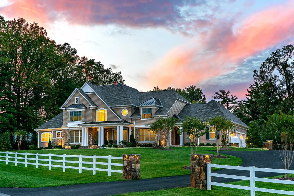 A large suburban house at sunset with landscaped lawn and trees, white fence, and illuminated windows, under a colorful sky with pink and purple clouds.