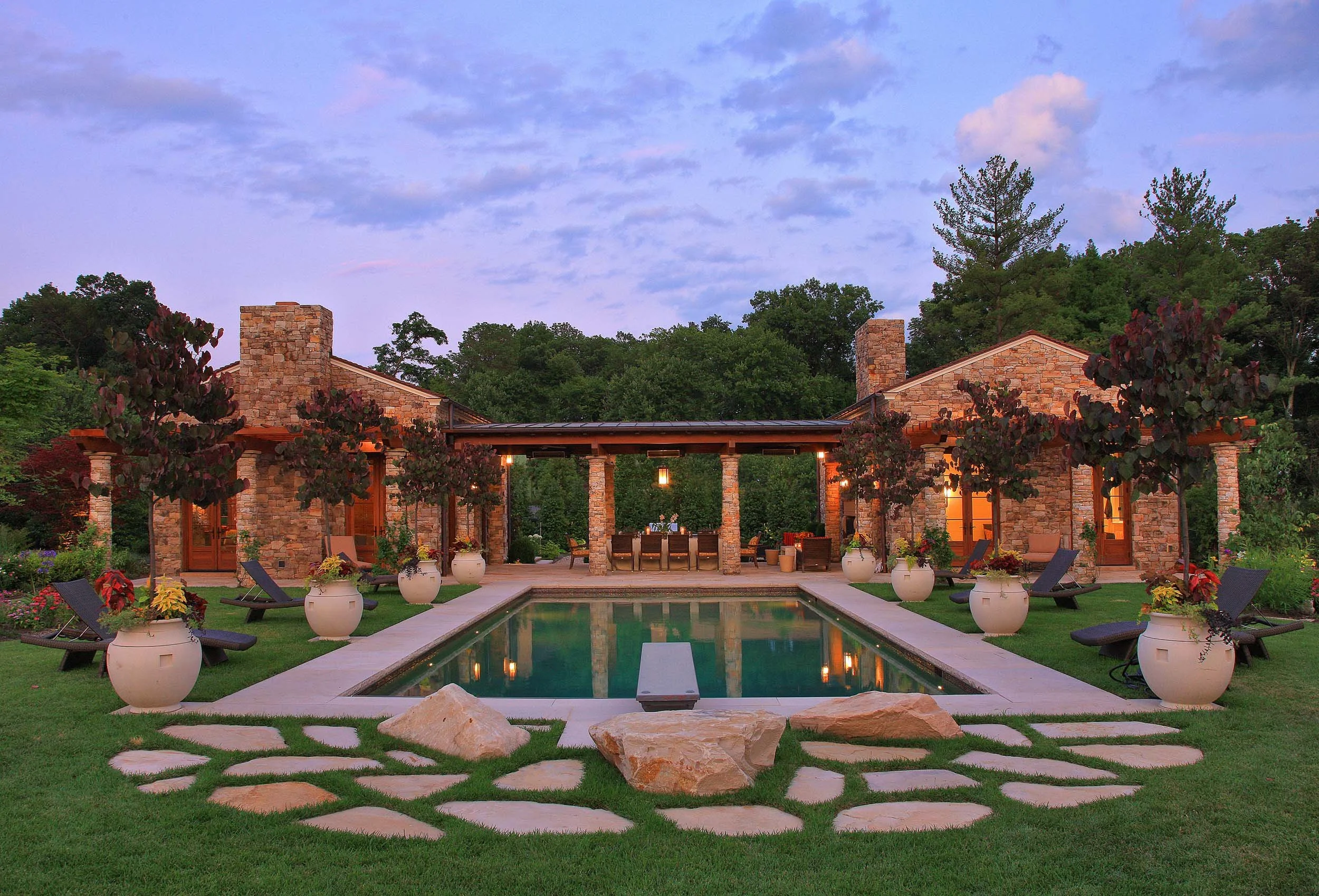 A backyard with a swimming pool, surrounded by potted plants, lounge chairs, and a stone house with a patio, trees, and a blue sky with clouds.