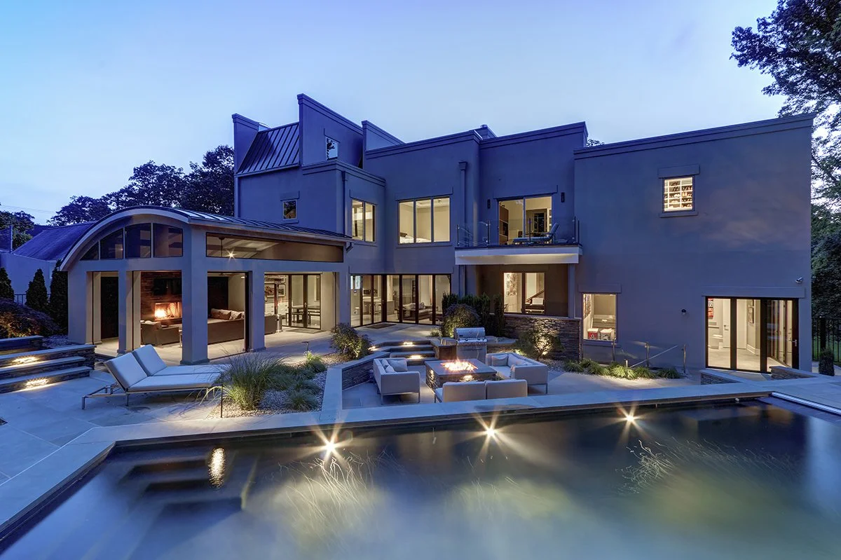 Modern multi-story house with large windows and a backyard with a swimming pool, outdoor seating area, and firepit, illuminated during dusk.