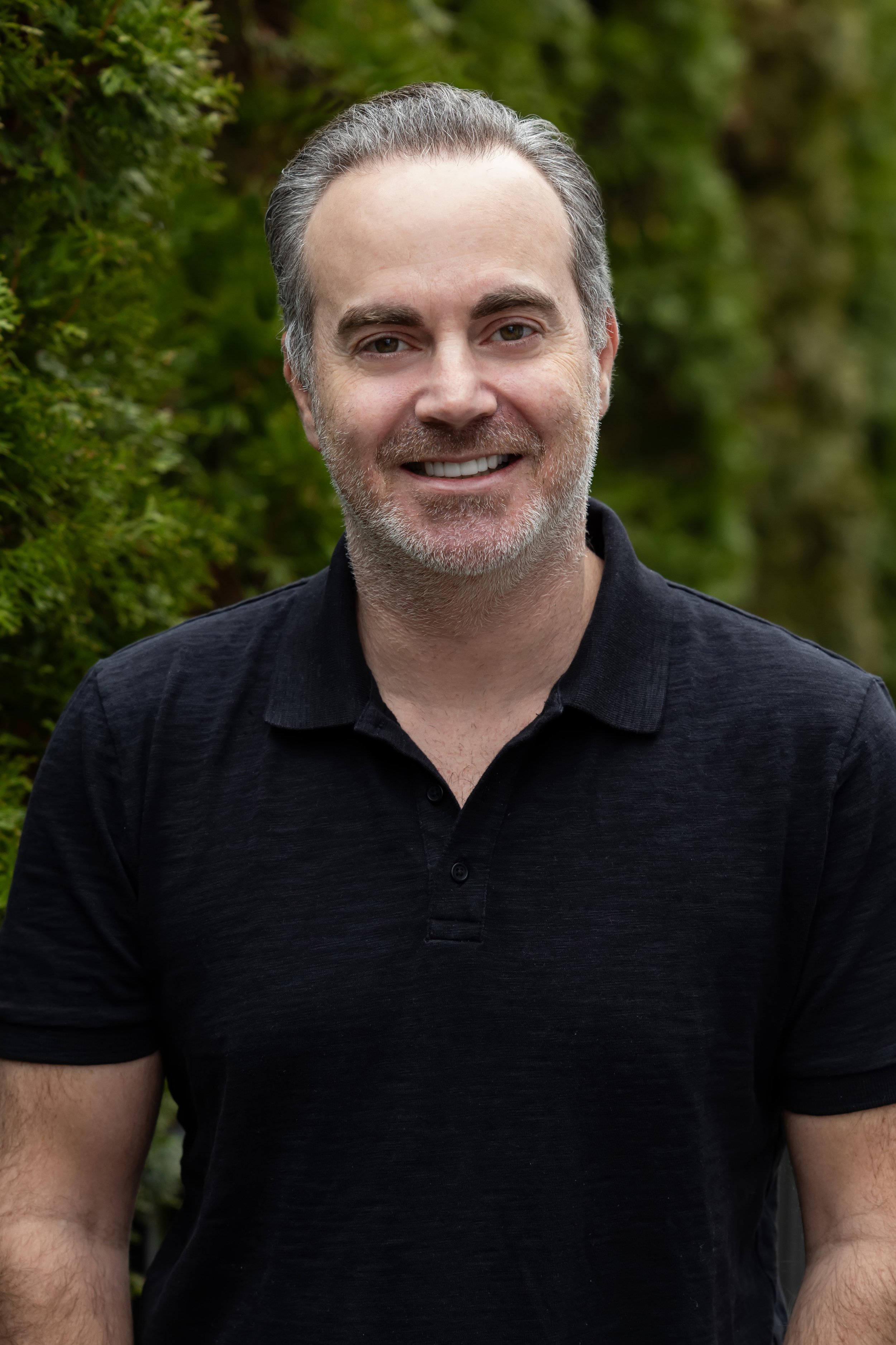 Close-up photo of a smiling man with gray beard and dark hair, wearing a black polo shirt, outdoors with green foliage in the background.