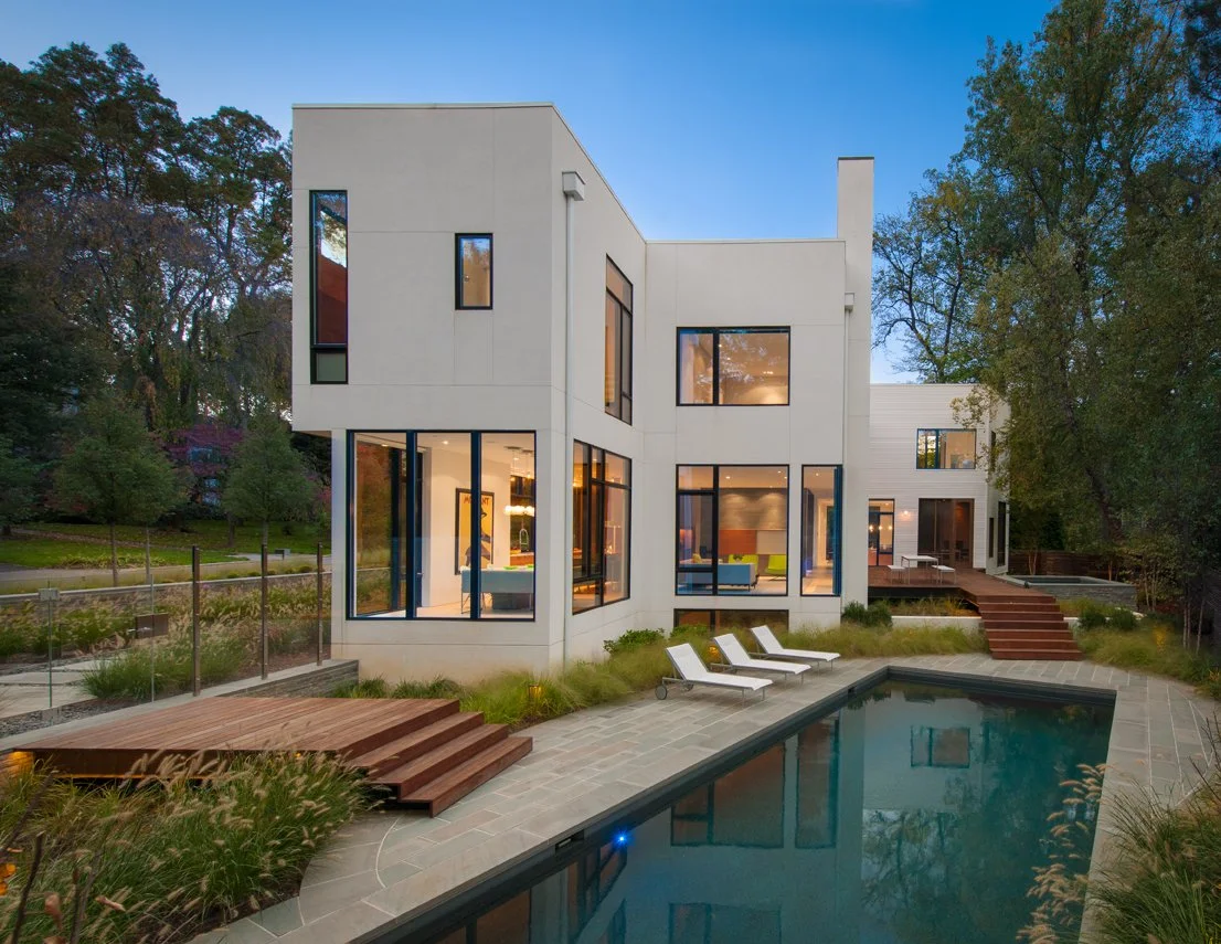 Modern white house with large windows, outdoor pool, and surrounding greenery at dusk.