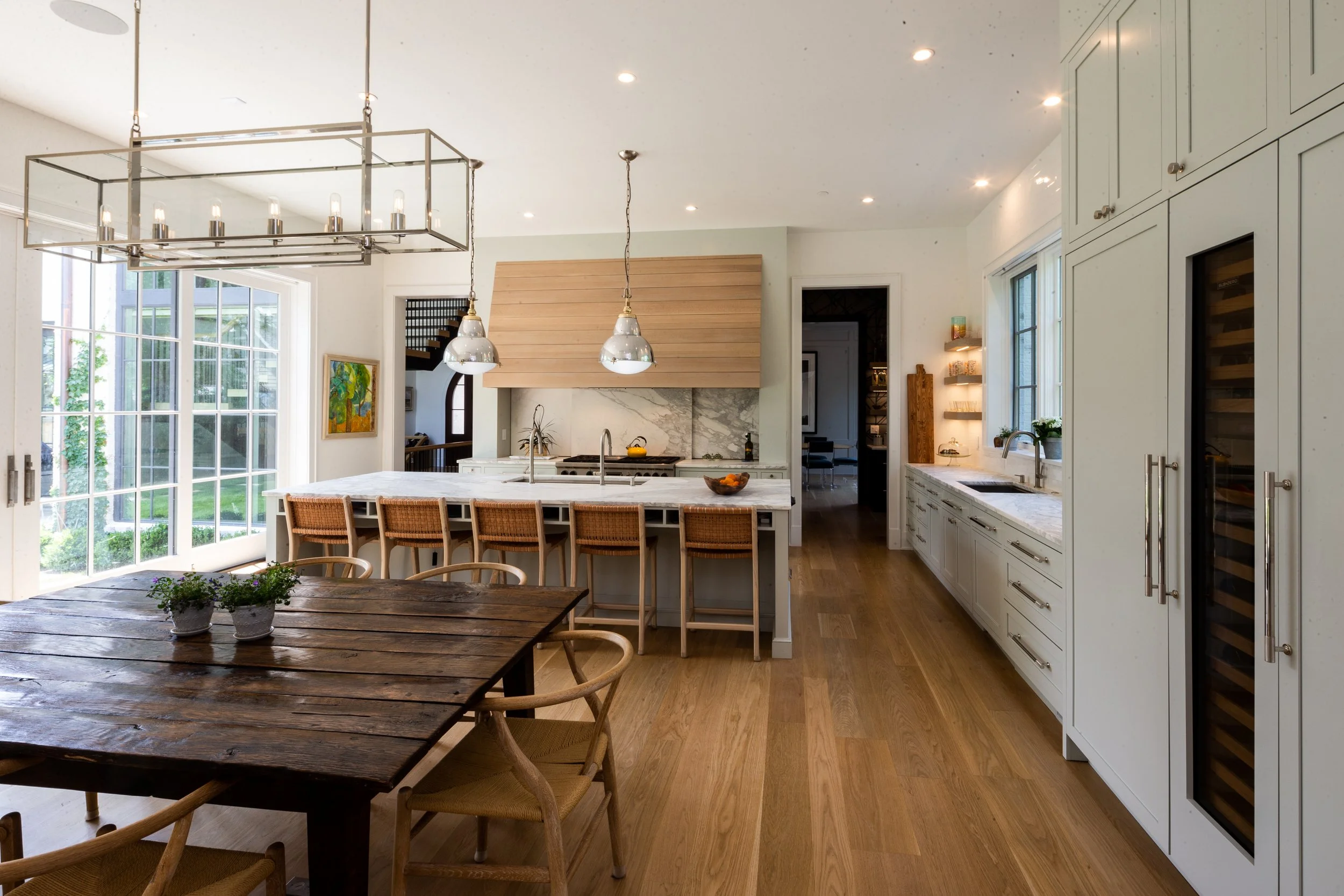 Modern kitchen with large island, white cabinetry, and wood accents. A wooden dining table with chairs is in the foreground. Large windows and glass doors allow natural light, and there are pendant and candelabra-style ceiling lights.