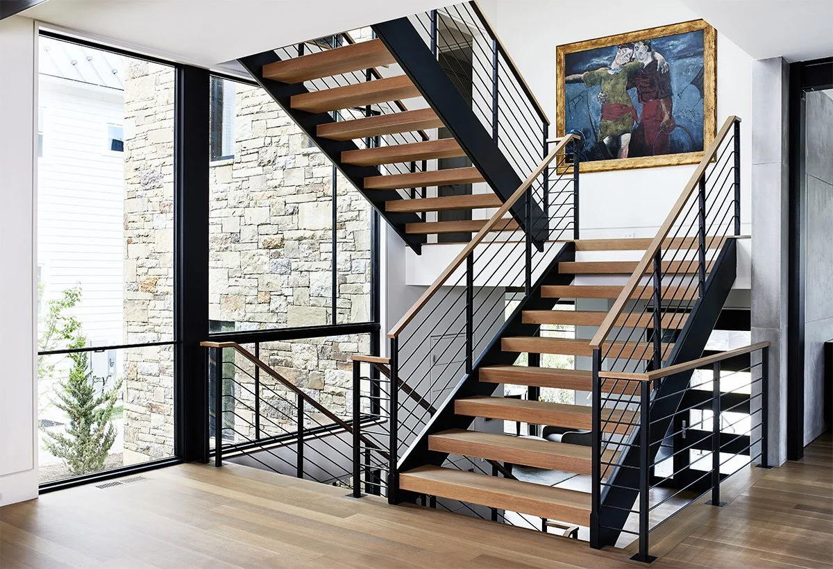 Modern staircase with wooden steps and black metal railings inside a home, next to large floor-to-ceiling windows, with a colorful abstract painting on the wall.