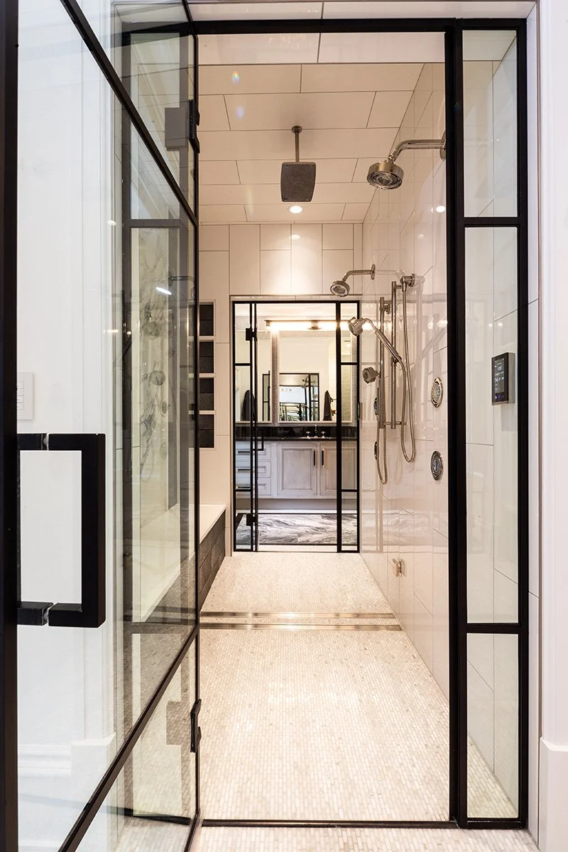 View of a modern walk-in shower with multiple showerheads, enclosed by glass doors and black metal framing, leading to a bathroom with white cabinetry and a mirror.
