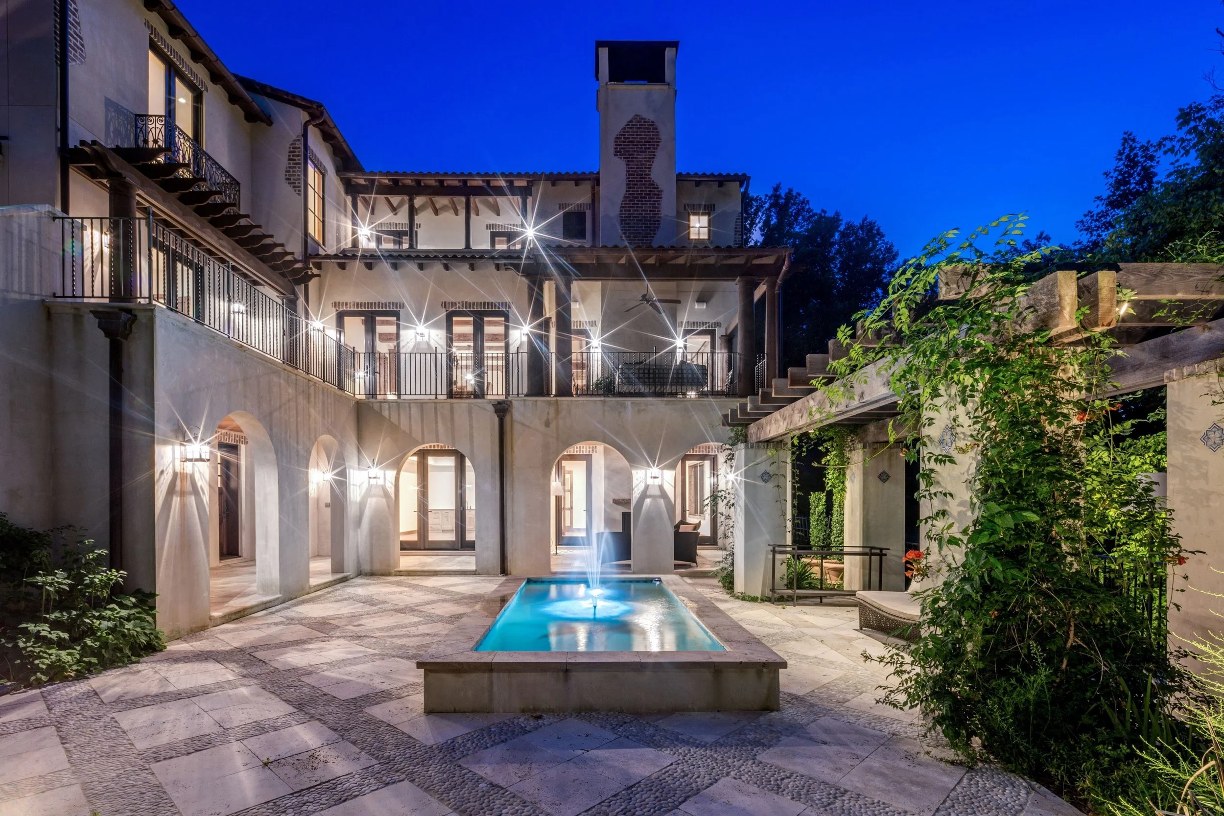 Night view of a Mediterranean-style villa with a rectangular swimming pool, outdoor lighting reflections, arched walkways, a chimney with cutout patterns, and lush greenery.