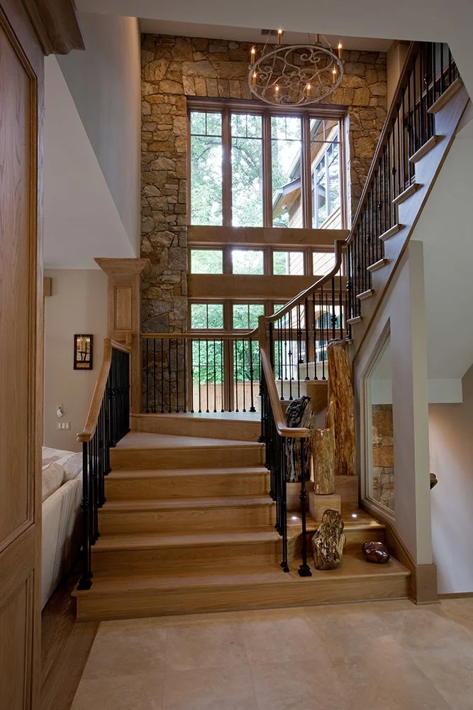 Interior view of a wooden staircase with black metal railings in a rustic home, featuring a stone accent wall, large windows, and a chandelier overhead.