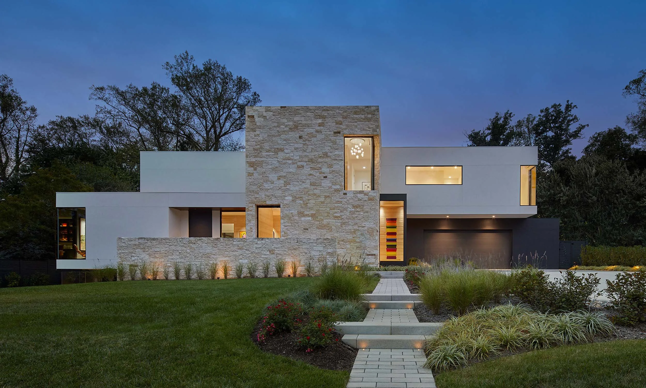 Modern house with stone and white exterior, large windows, landscaped yard, and pathway leading to the entrance during dusk.