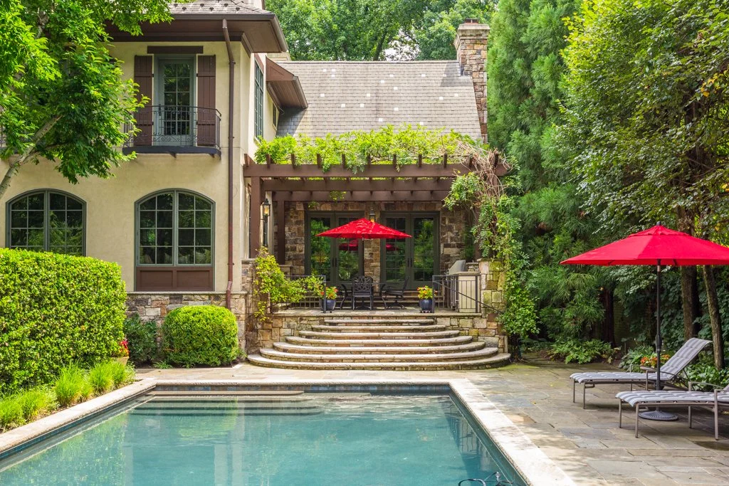 A backyard with a swimming pool, surrounding green bushes, and a house with large windows and a terrace with red umbrellas and outdoor furniture.