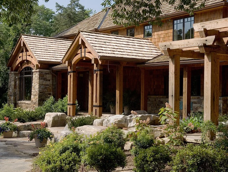Front view of a two-story house with stone and wood exterior, a gabled roof, and a porch with wooden columns, surrounded by landscaped garden with plants and rocks.