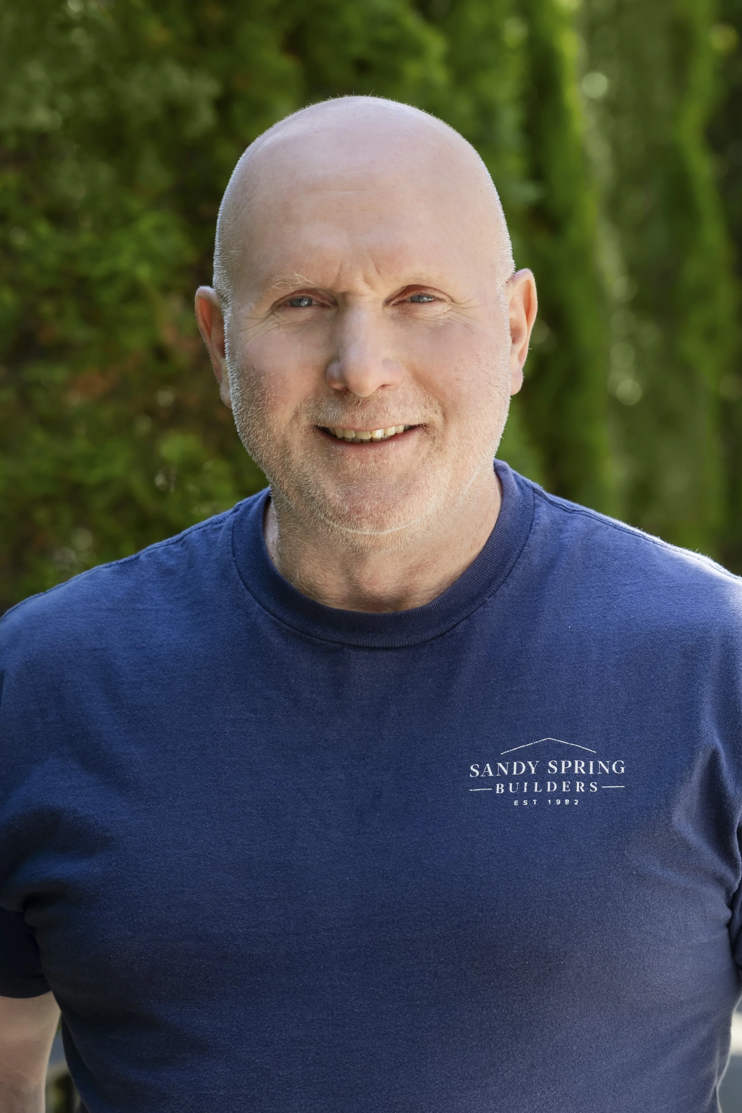 Close-up of a middle-aged man with a shaved head, wearing a navy blue t-shirt with the logo 'Sandy Spring Builders, Est 1982' printed on it, smiling outdoors with a blurred green foliage background.