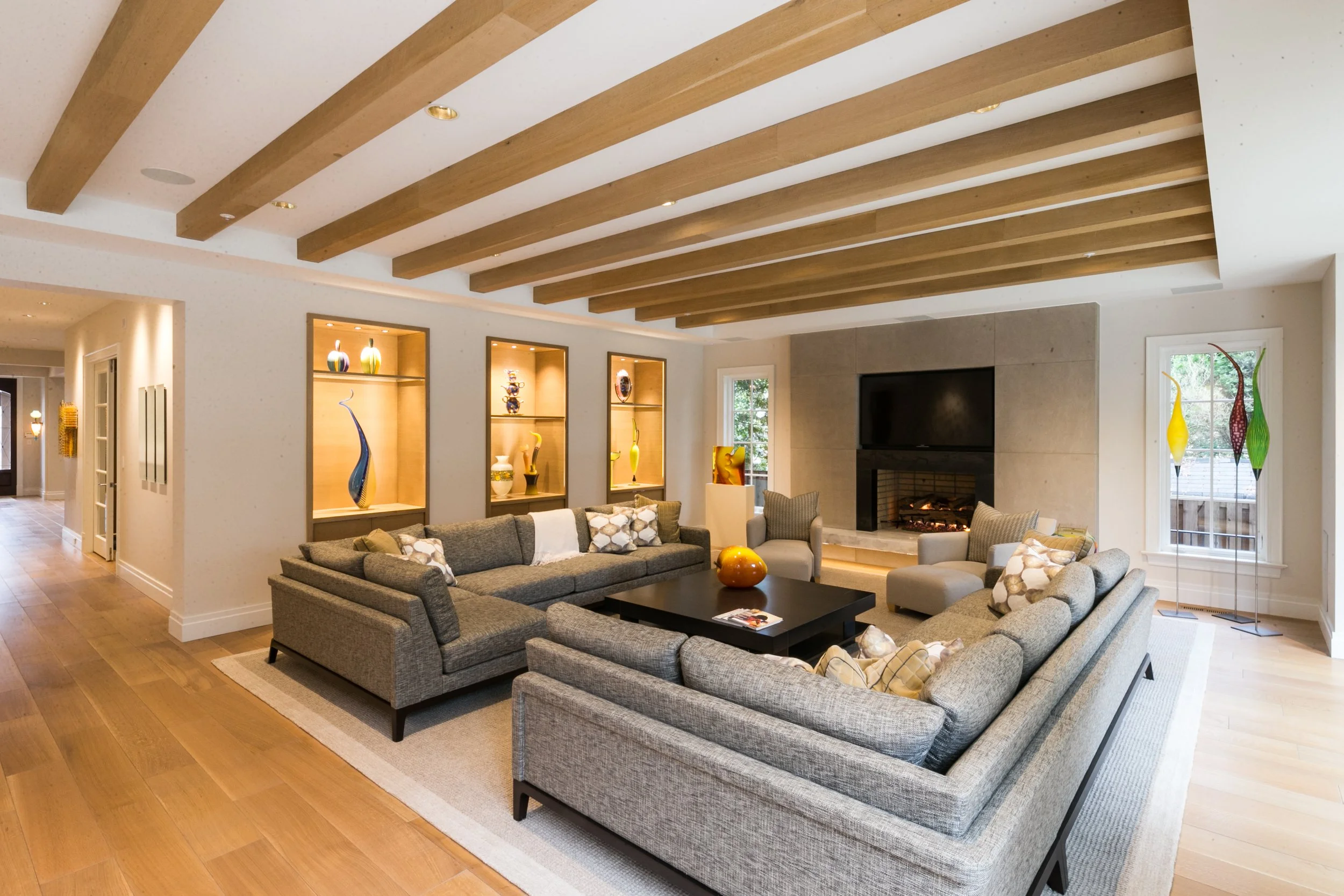 Living room with wooden beam ceiling, gray sofas, a black coffee table, decorative vases, a fireplace, and windows with greenery outside.