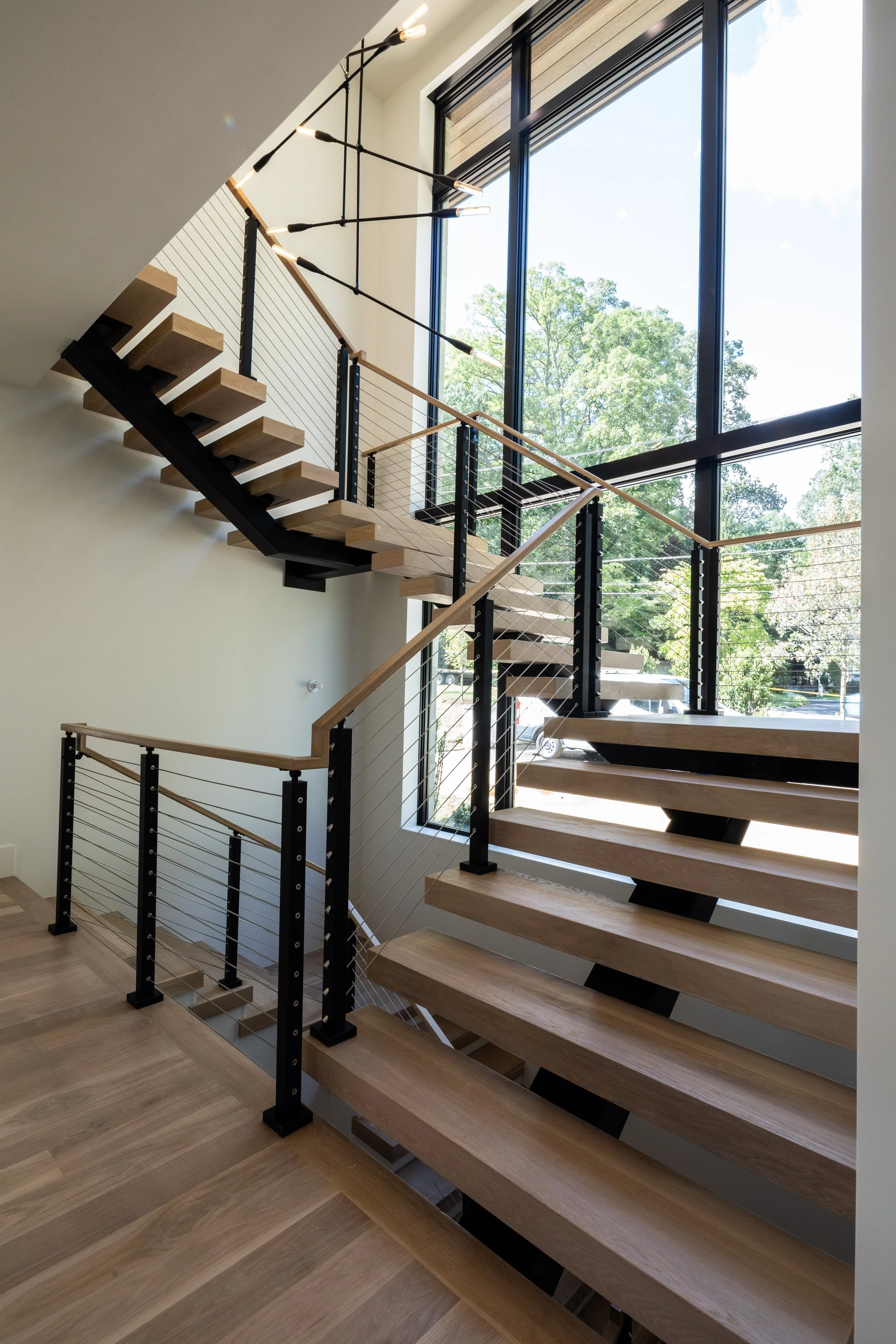 Modern staircase with wooden steps, black metal supports, and cable railing, next to large floor-to-ceiling windows showing trees outside.
