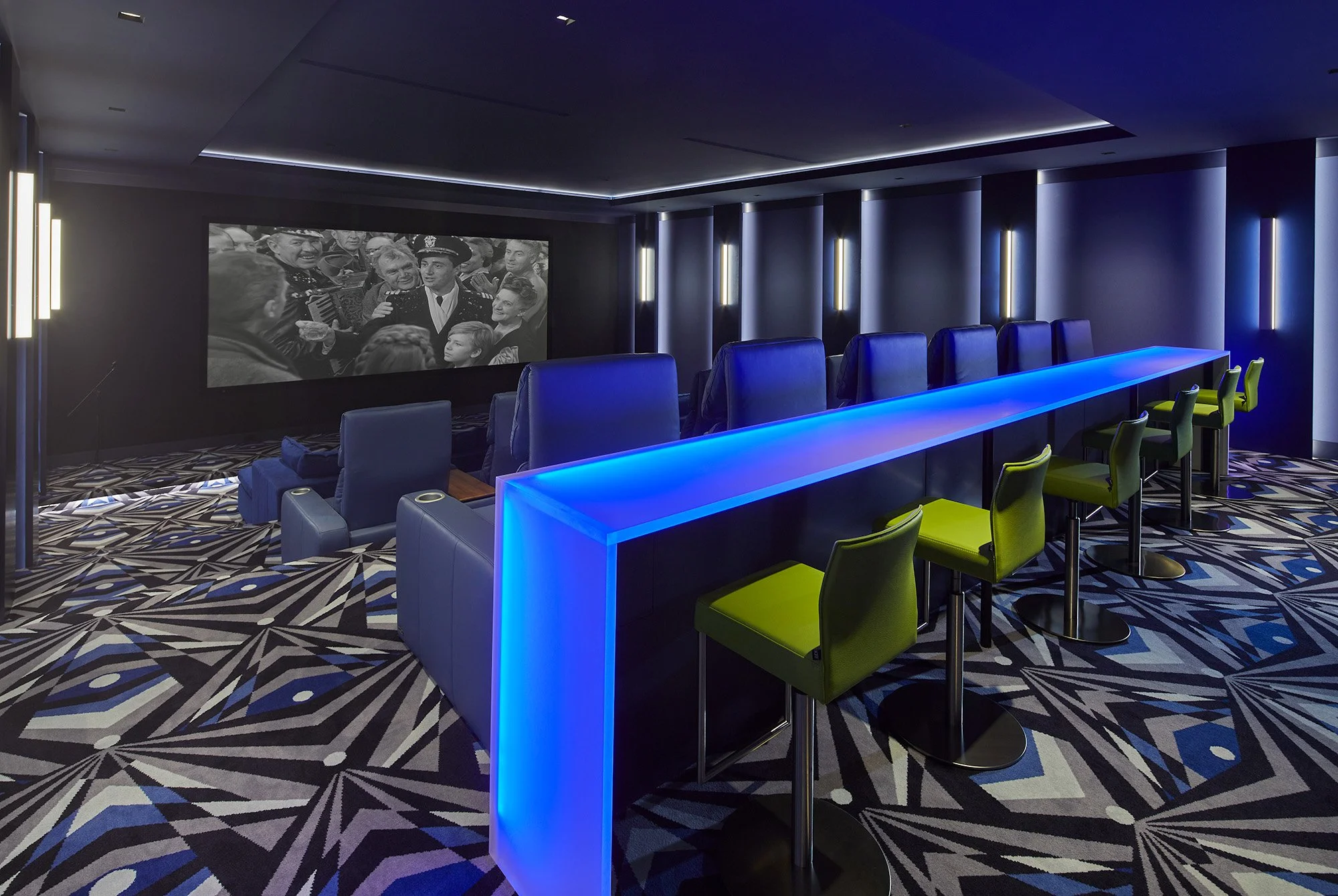 A modern home theater room with a large screen displaying a black-and-white movie scene. The room features blue LED lighting along the ceiling and a long blue-lit bar with green chairs. There's a patterned carpet and plush blue seating in front of th