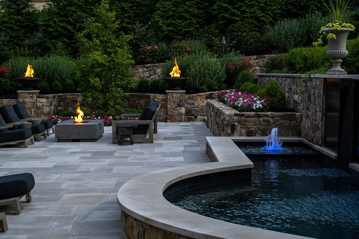 Outdoor patio with a pool, fire pits, and seating area surrounded by lush green garden and stone walls.
