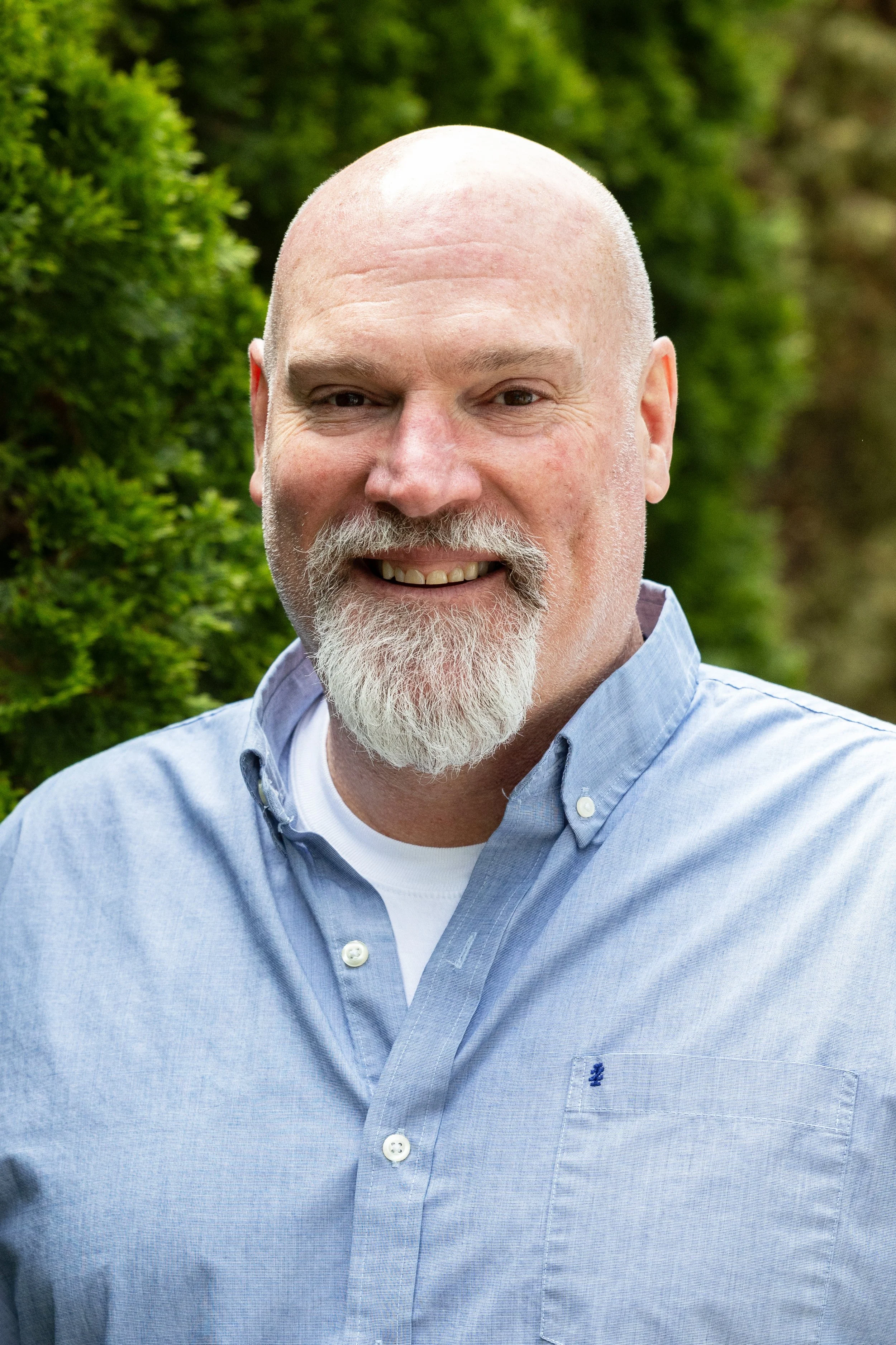 Close-up of a smiling middle-aged man with a bald head and a white beard, wearing a light blue collared shirt, outdoors with greenery in the background.