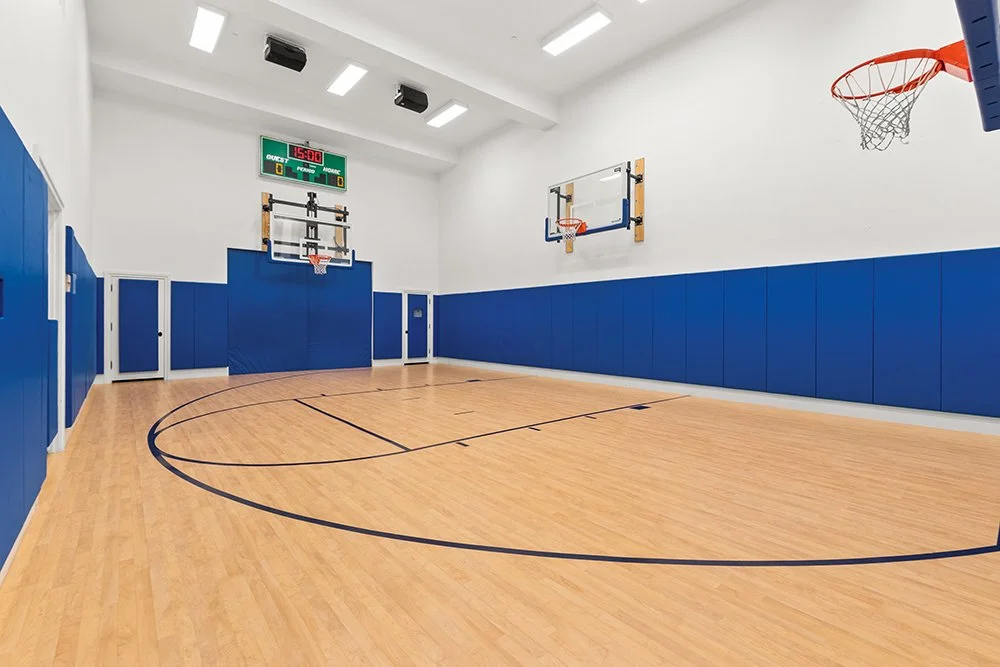 Empty indoor basketball court with wooden flooring, blue padded walls, two basketball hoops, and a digital scoreboard.
