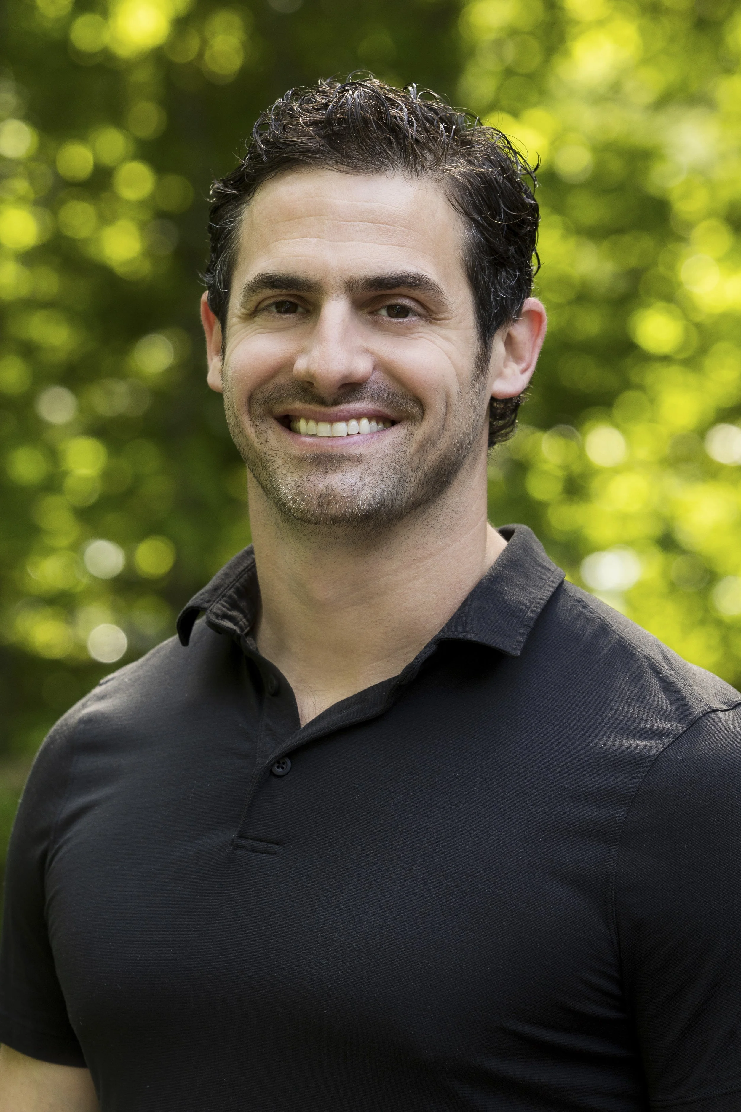 Close-up of a smiling man with dark curly hair, wearing a black collared shirt, standing outdoors with blurred green foliage in the background.