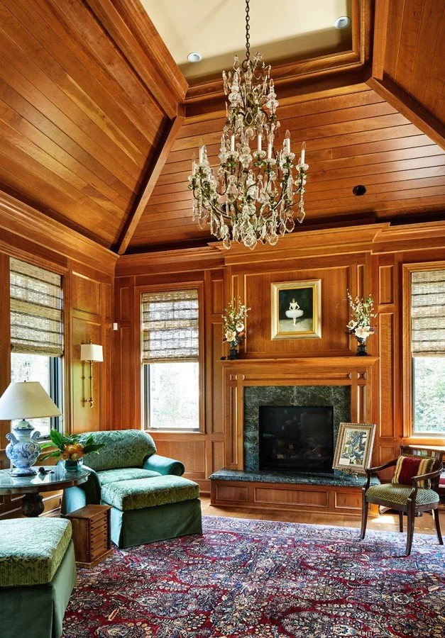 Wood-paneled living room with a chandelier, green sofa, fireplace, framed artwork, and large windows with window treatments.