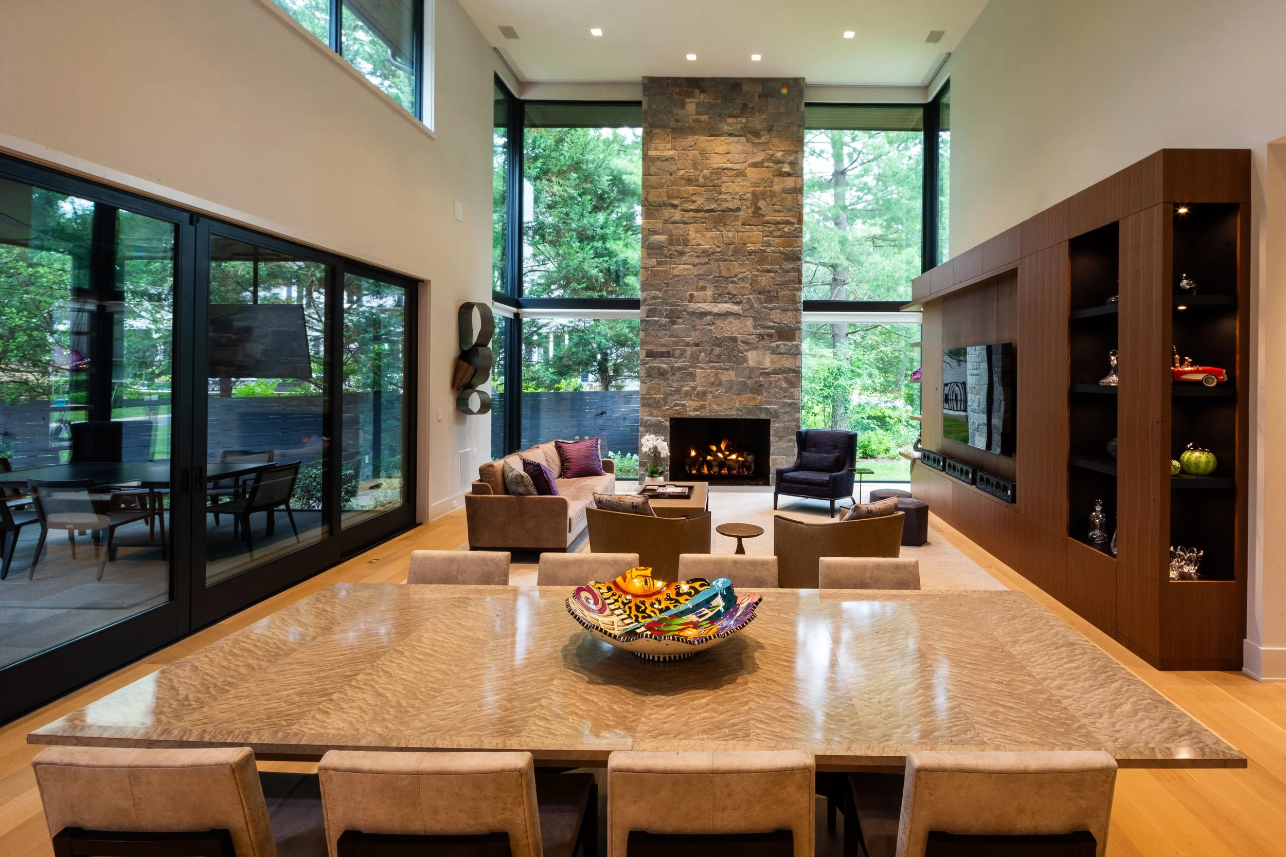 Modern living room with large windows, a stone fireplace, wooden cabinetry, and a dining table with chairs in the foreground.