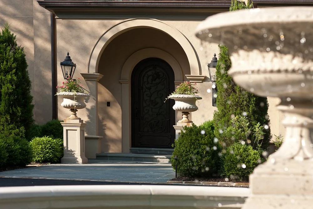 Elegant front entrance of a house with an arched doorway, black wrought iron door, flower planters on pedestals, lush green bushes, and a fountain.