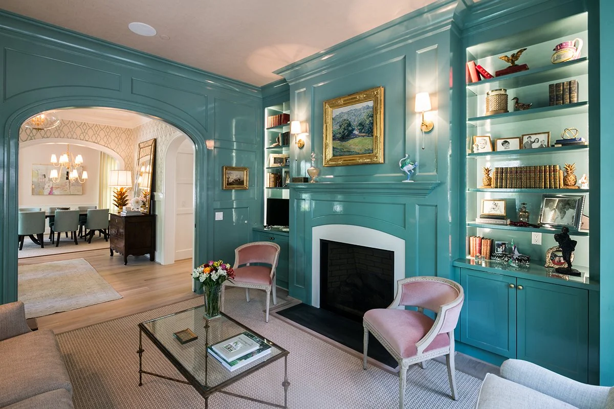 Living room with teal walls, built-in bookshelves, a fireplace, pink chairs, a glass coffee table with books and a flower vase, and a view into a dining area with a chandelier and green chairs.