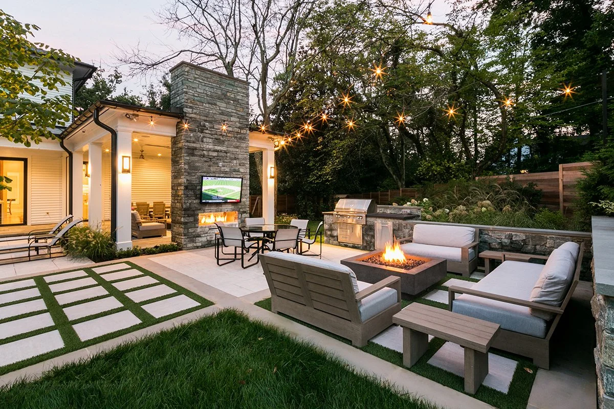 An outdoor backyard patio with lounge furniture, a fire pit, string lights, a built-in fireplace with a television, and a barbecue grill, surrounded by trees and a wooden fence.