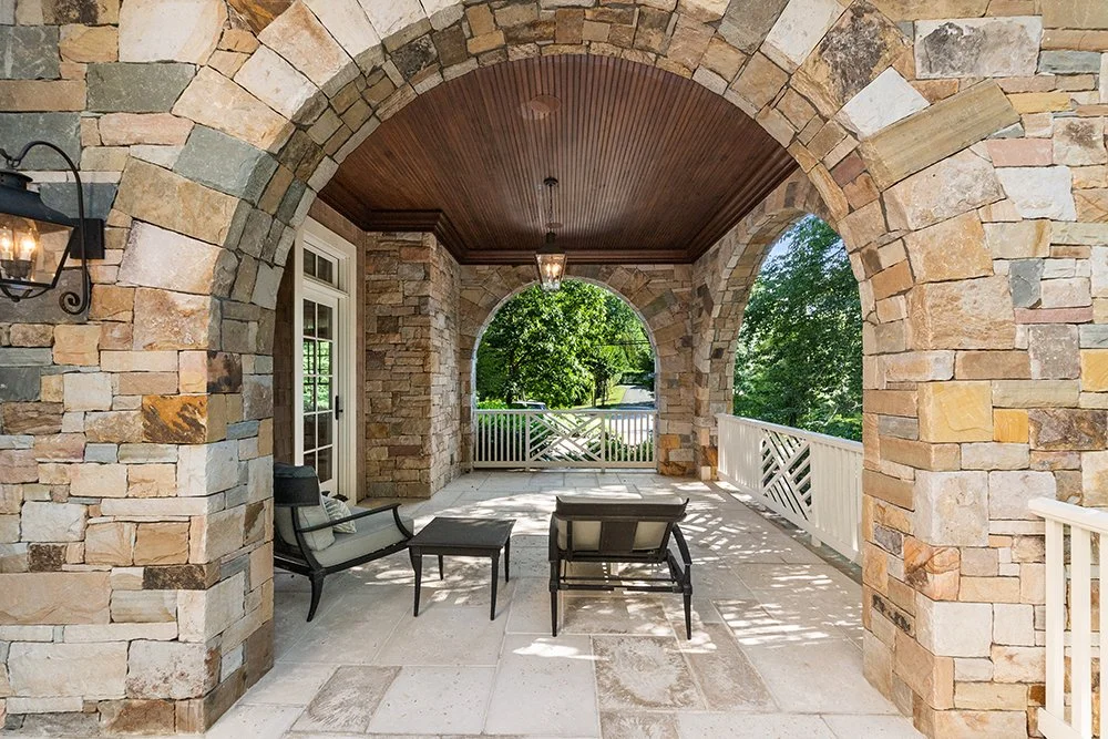 Outdoor covered patio area with stone archways, wood-paneled ceiling, patio furniture, and a view of green trees and a white railing.