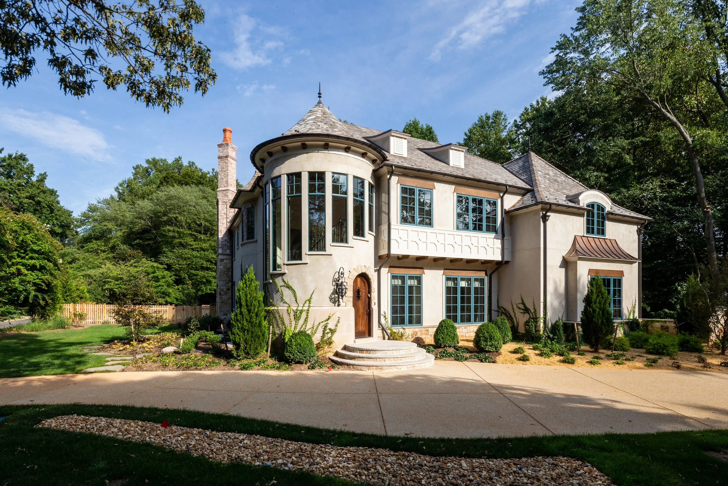 A large, elegant house with a rounded turret and multiple windows, surrounded by greenery and trees, under a clear blue sky.